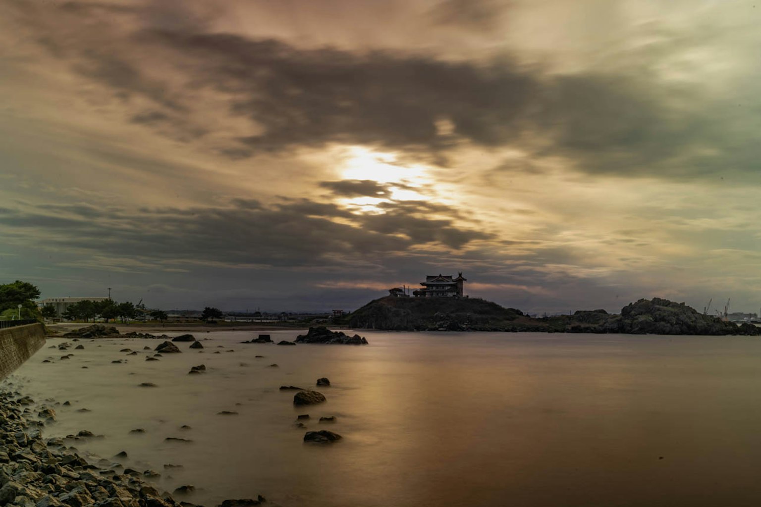 ทิวทัศน์ทะเลที่เงียบสงบ มีชายหาดหินและพระอาทิตย์ตกอยู่เบื้องหลัง