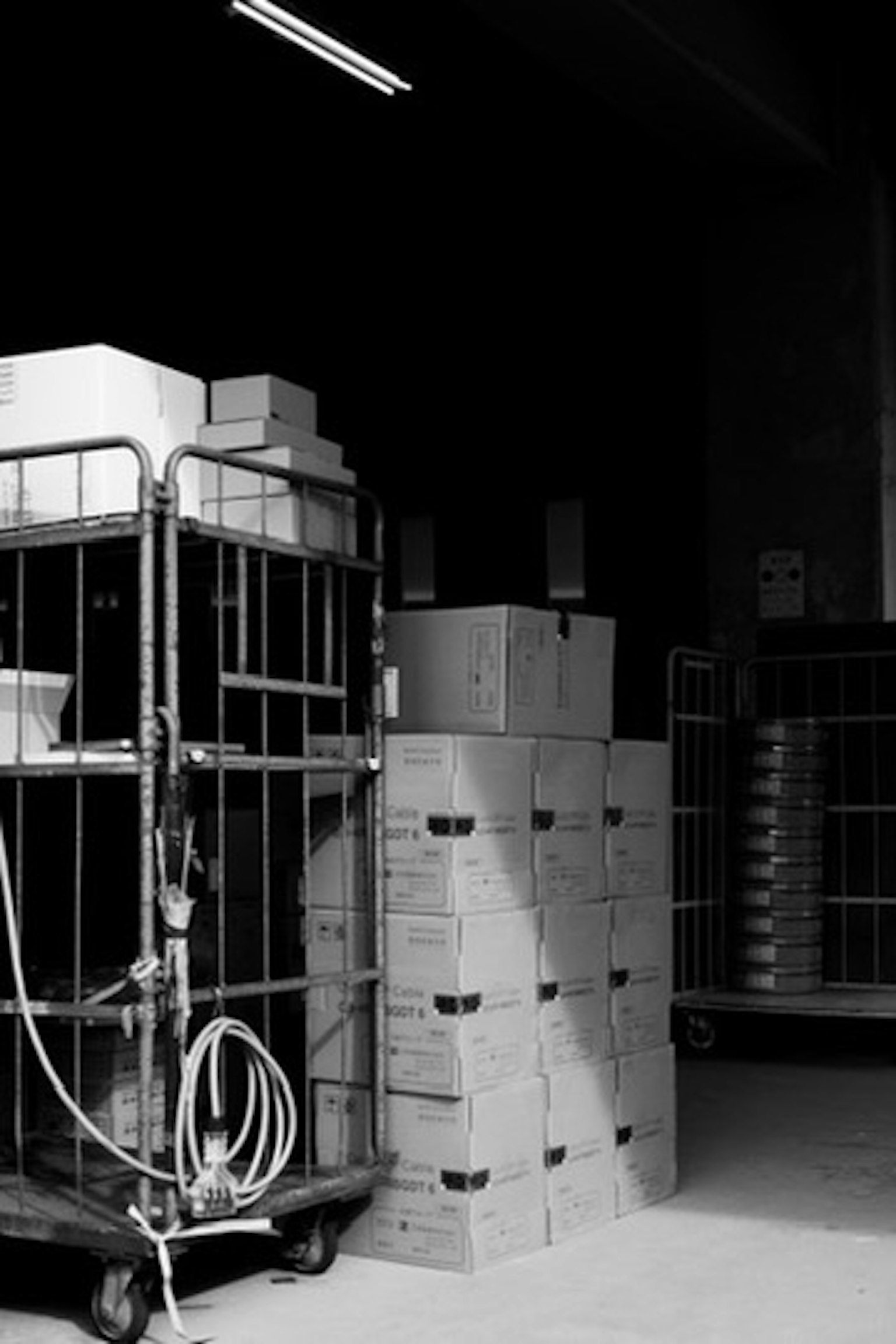 Interior en blanco y negro de un almacén con cajas de cartón apiladas y un carrito de transporte