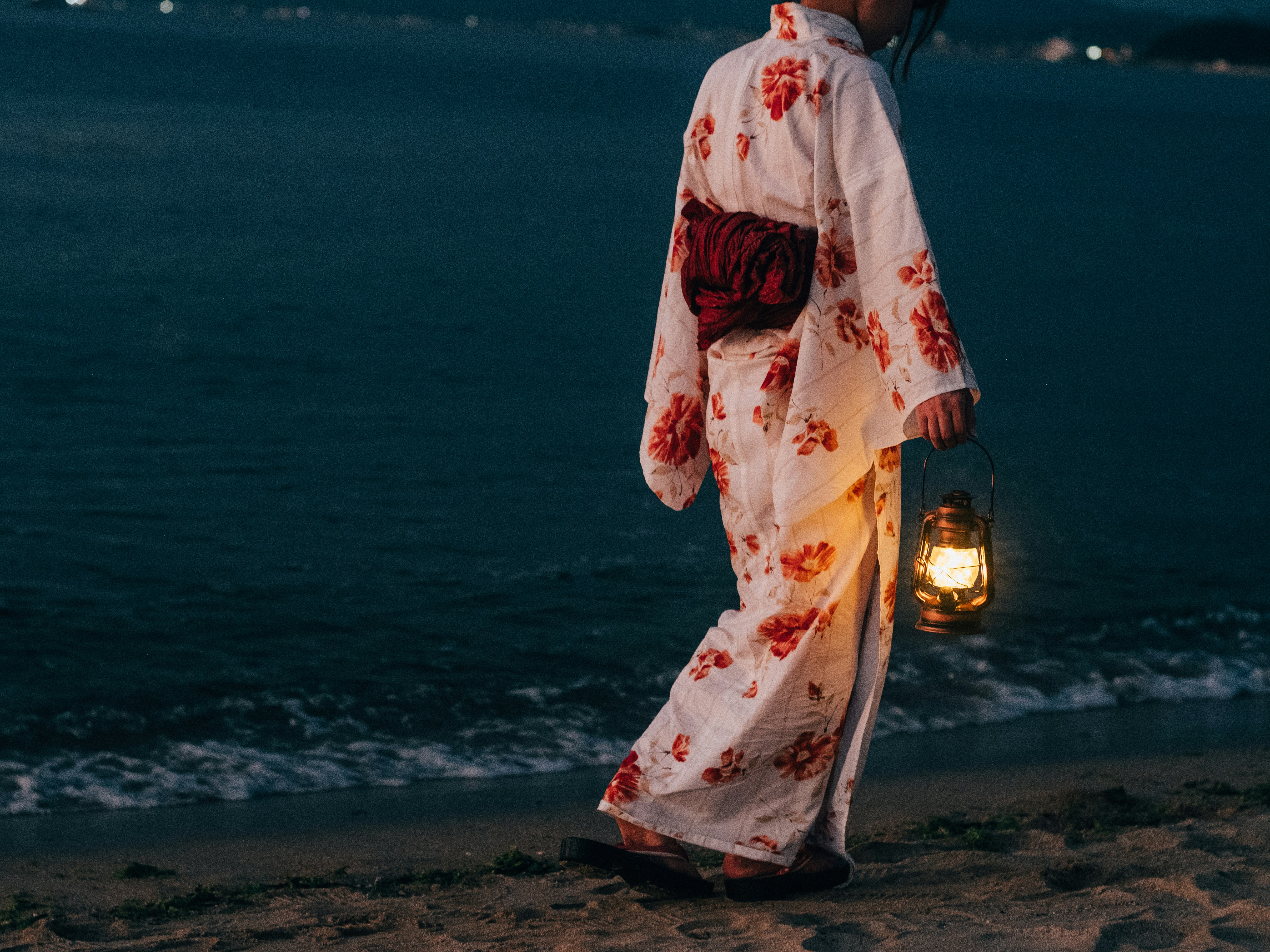 Femme en kimono fleuri marchant sur la plage avec une lanterne