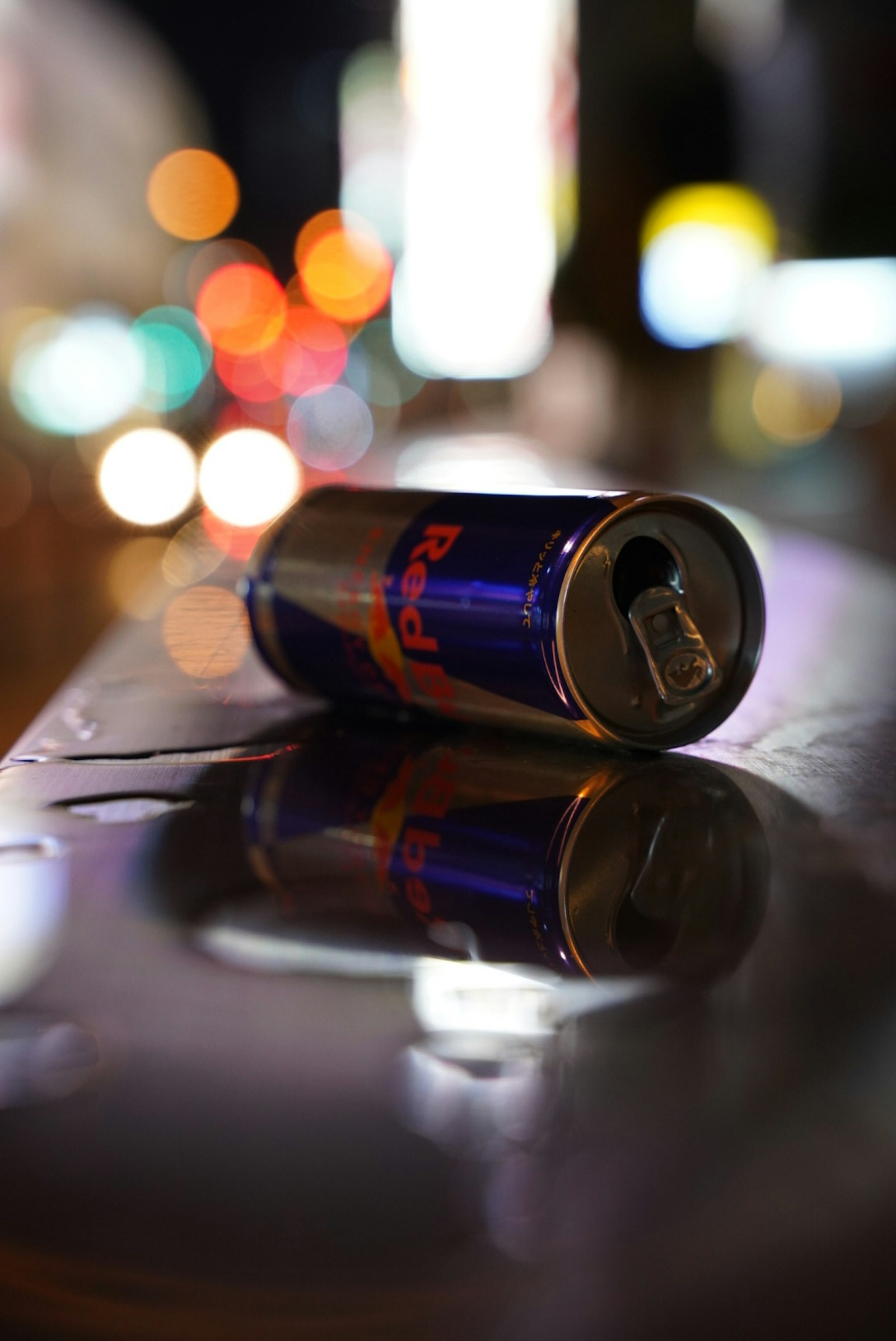A discarded Red Bull can lying on a shiny surface with blurred city lights in the background