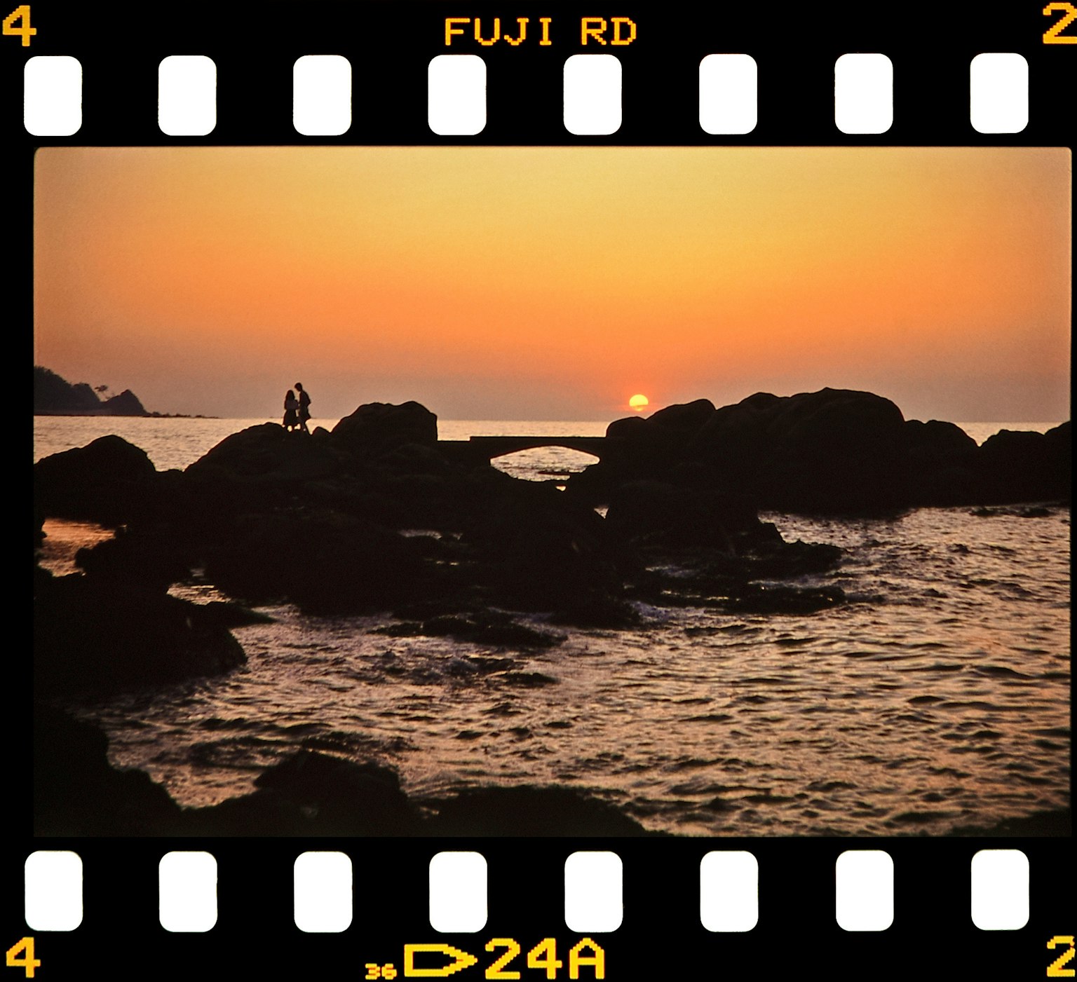 Siluet pasangan berdiri di atas batu saat matahari terbenam di tepi laut