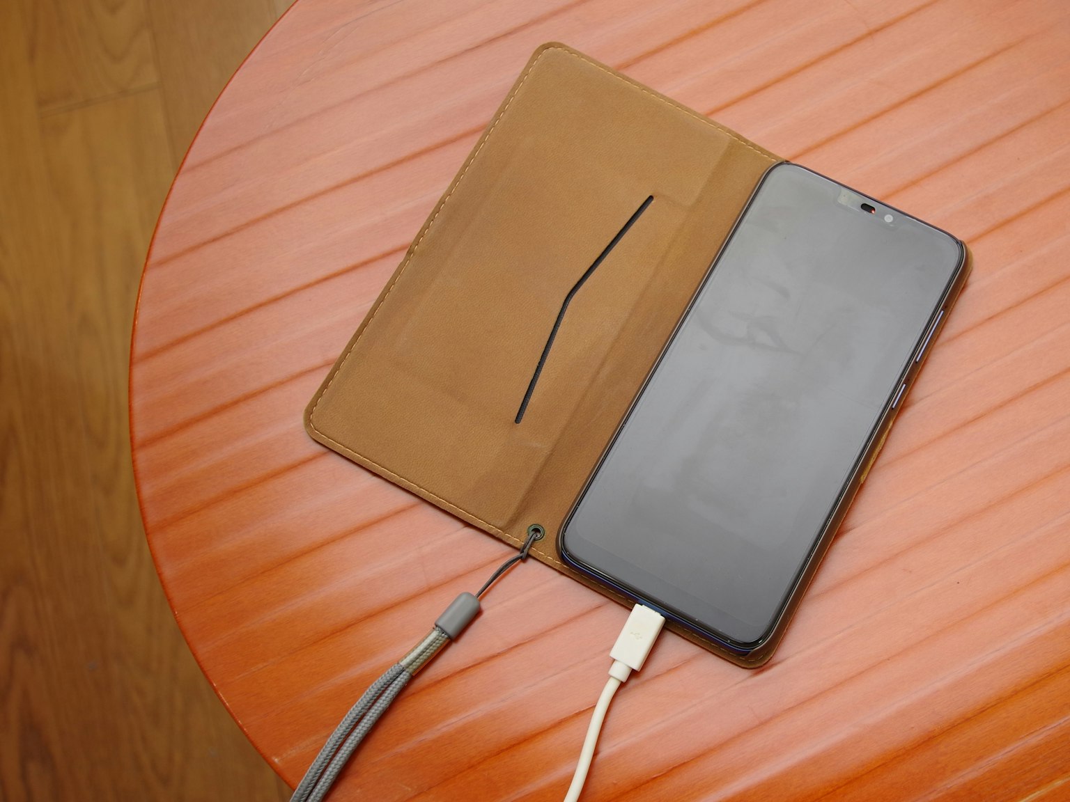 Smartphone dalam case kulit cokelat yang sedang diisi daya di atas meja kayu