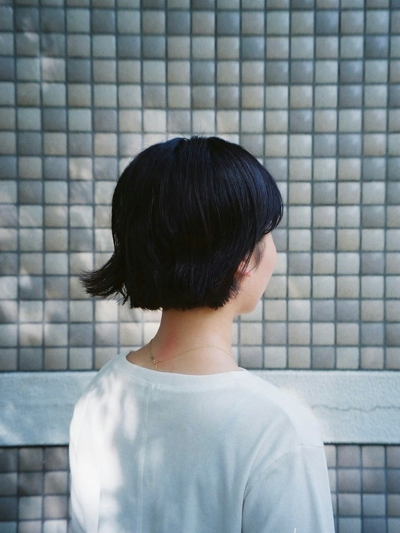 背中を向けた黒髪の女性がタイルの壁の前に立っている