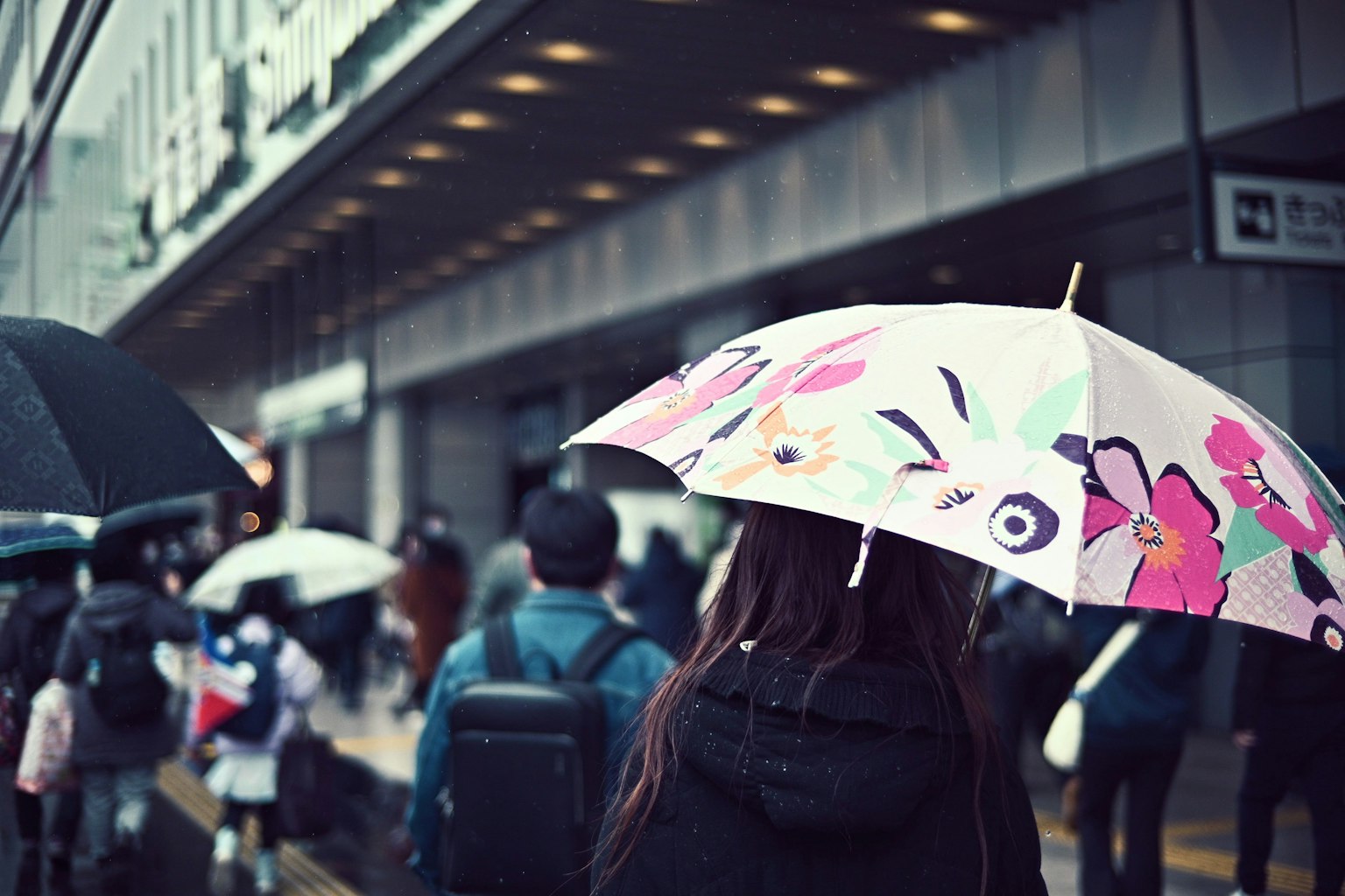 Một người phụ nữ cầm ô đầy màu sắc đi dưới mưa với đám đông người và phong cảnh đô thị ở phía sau