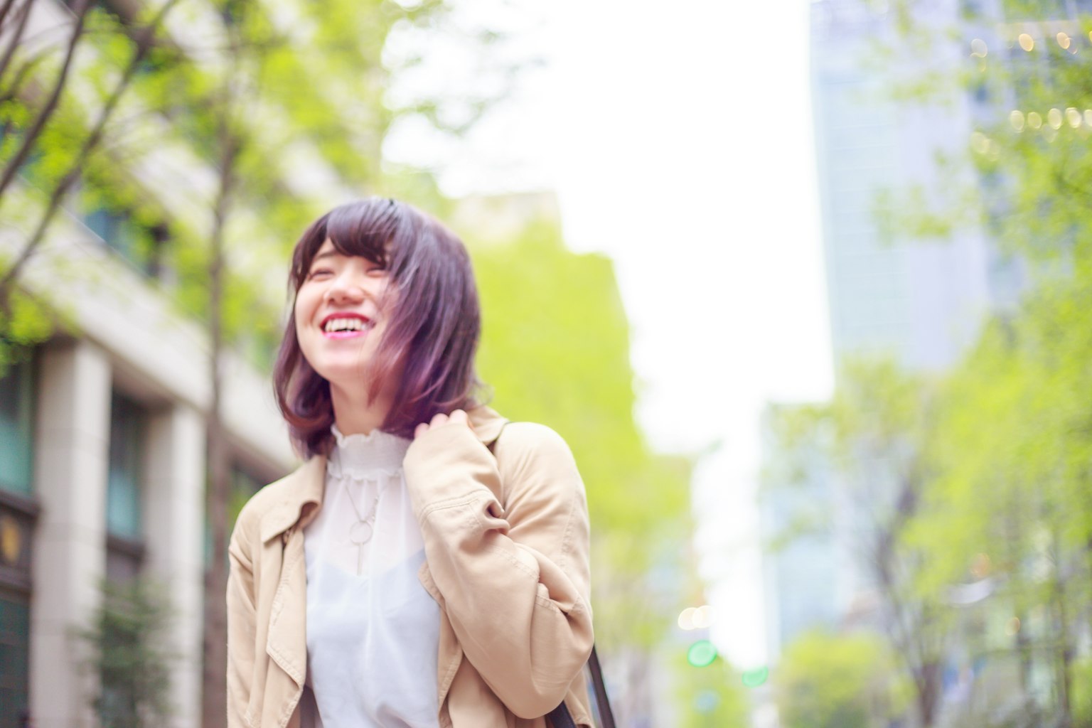 A young woman smiling in the street surrounded by green trees and tall buildings