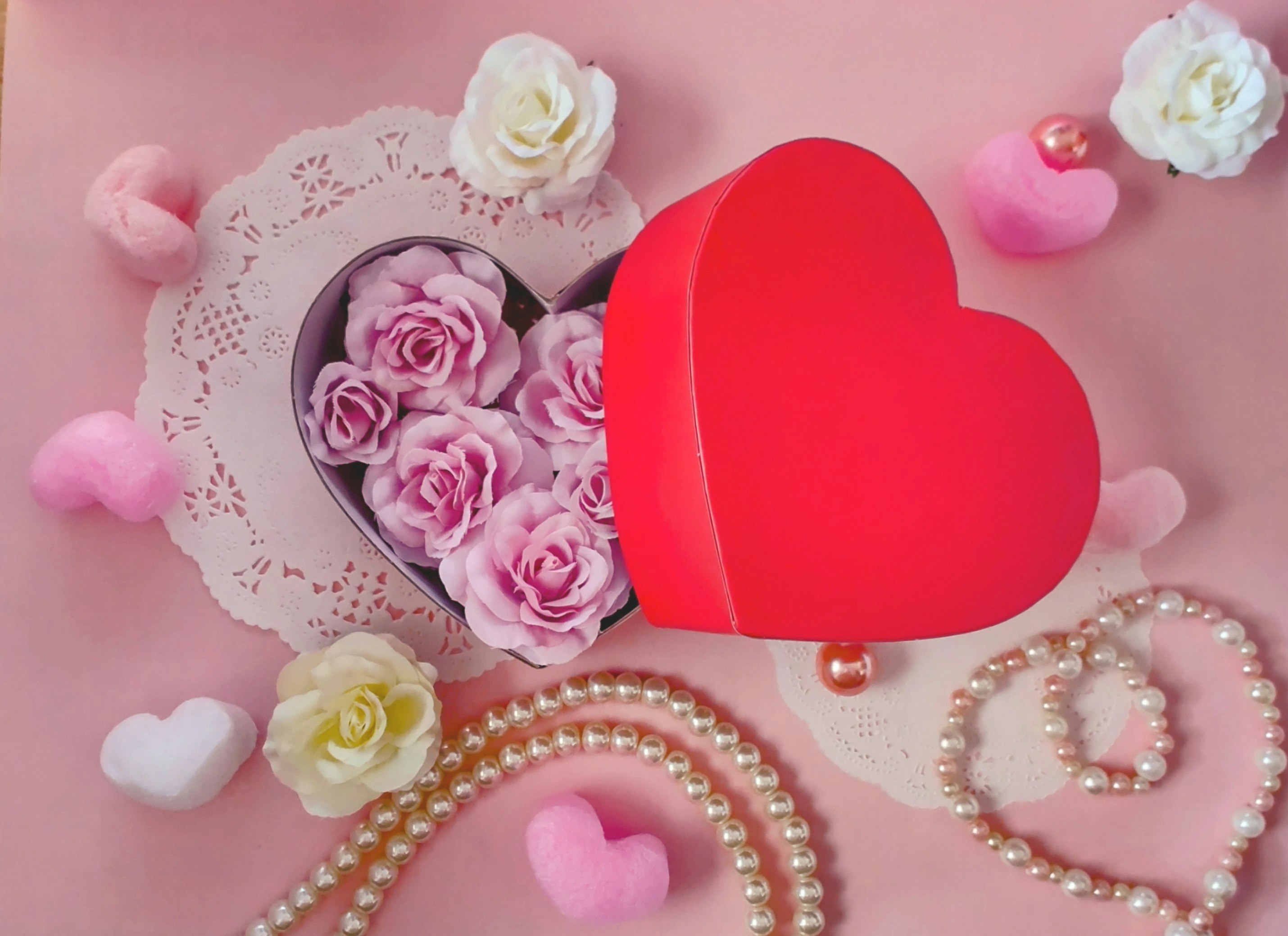 Valentine-style flat lay with a red heart, pearls, flowers, and lace paper on pink background