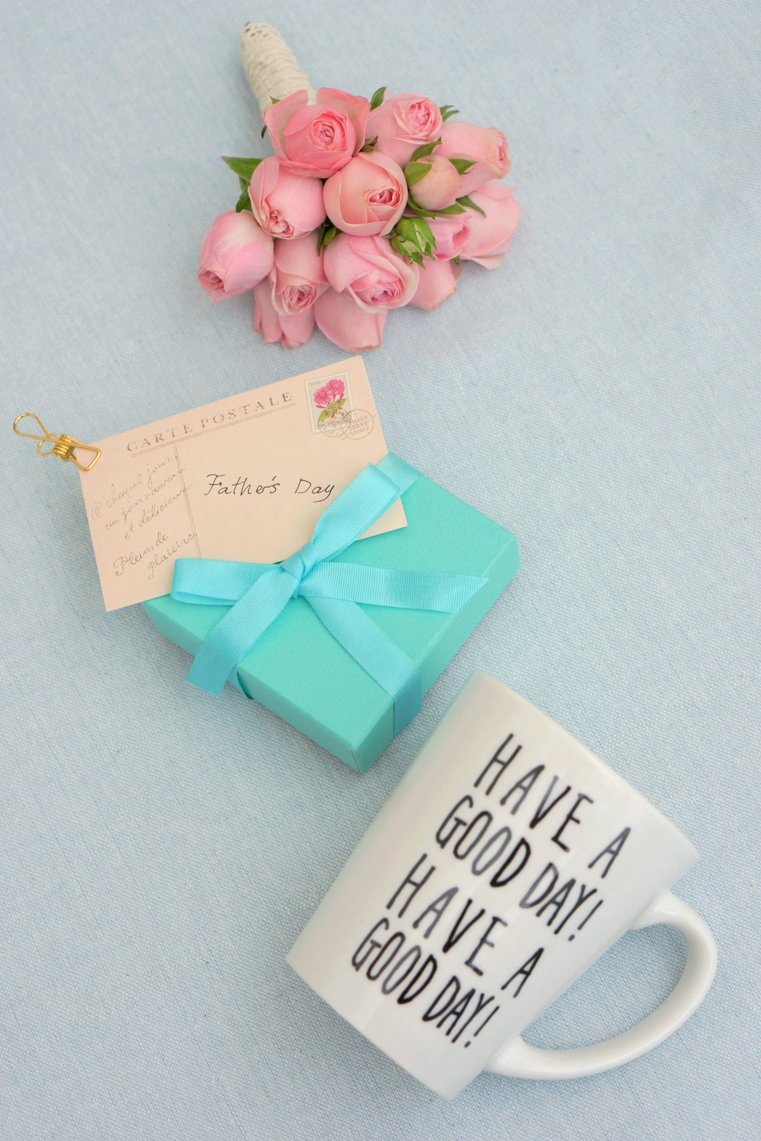 A pink flower bouquet, a turquoise wrapped gift with a tag, and a white mug with the message Have a Good Day on a blue background