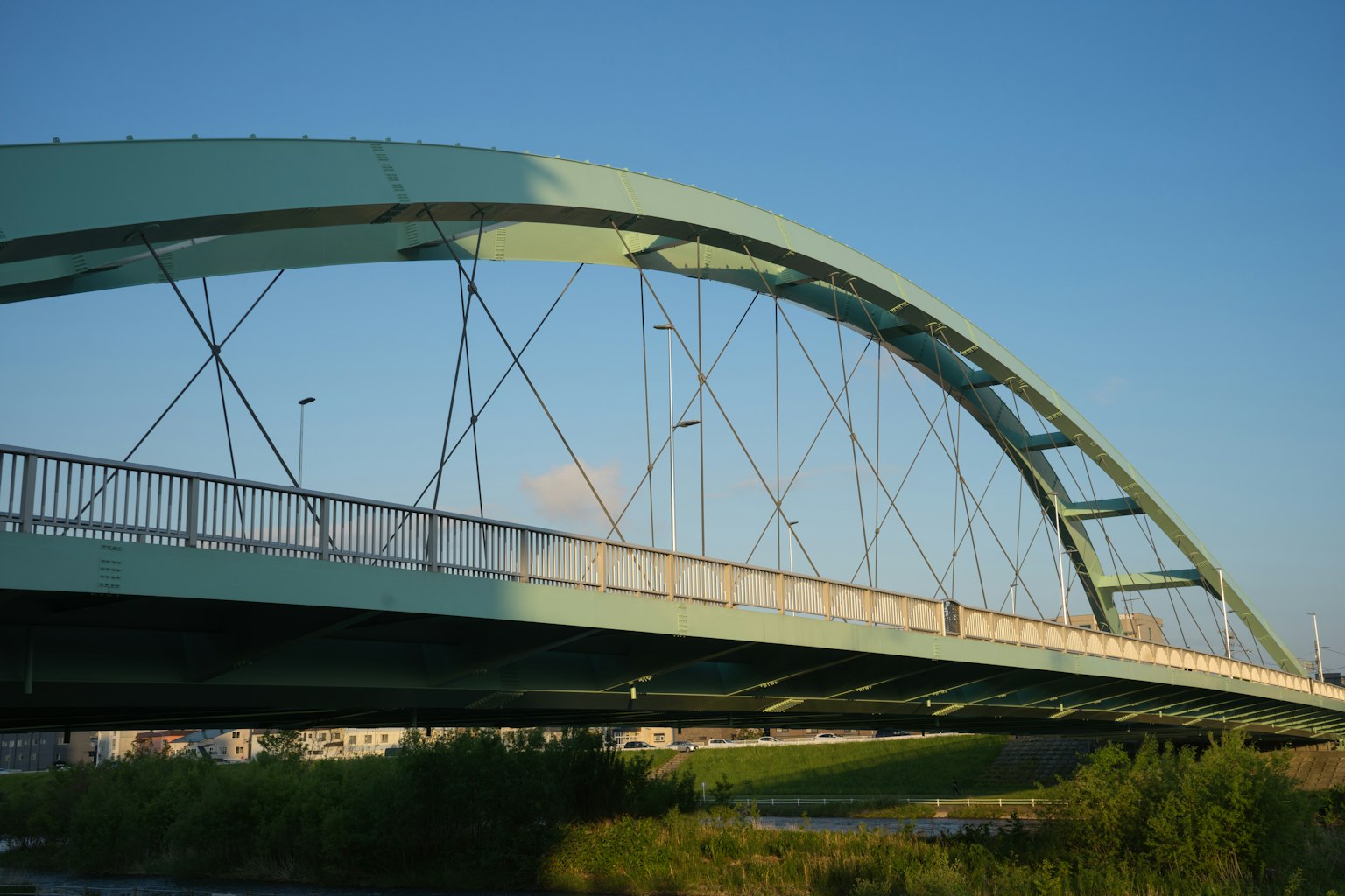 Jembatan lengkung berwarna teal melintasi sungai di bawah langit biru yang cerah