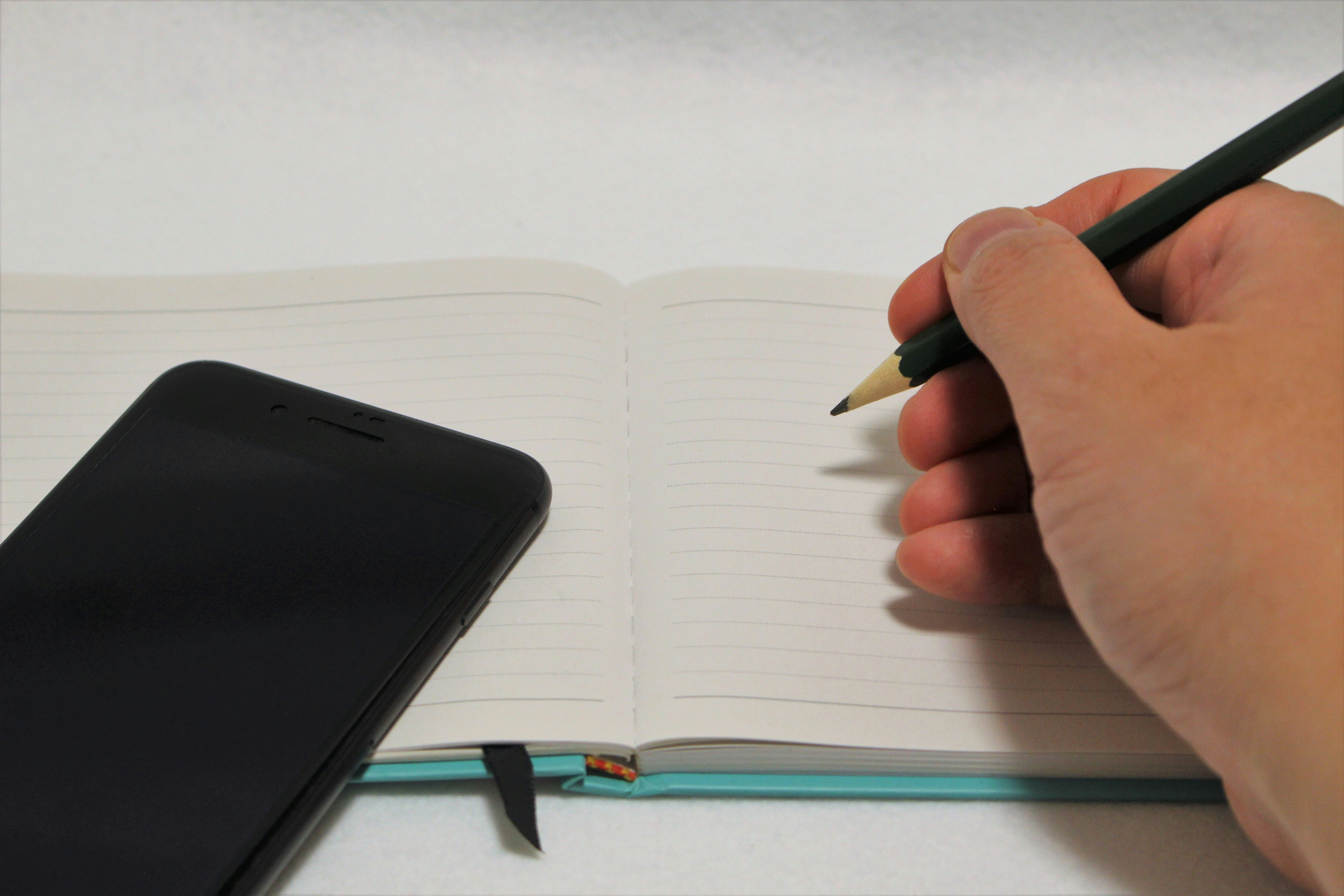 hand writing with a pencil in a notebook beside a smartphone