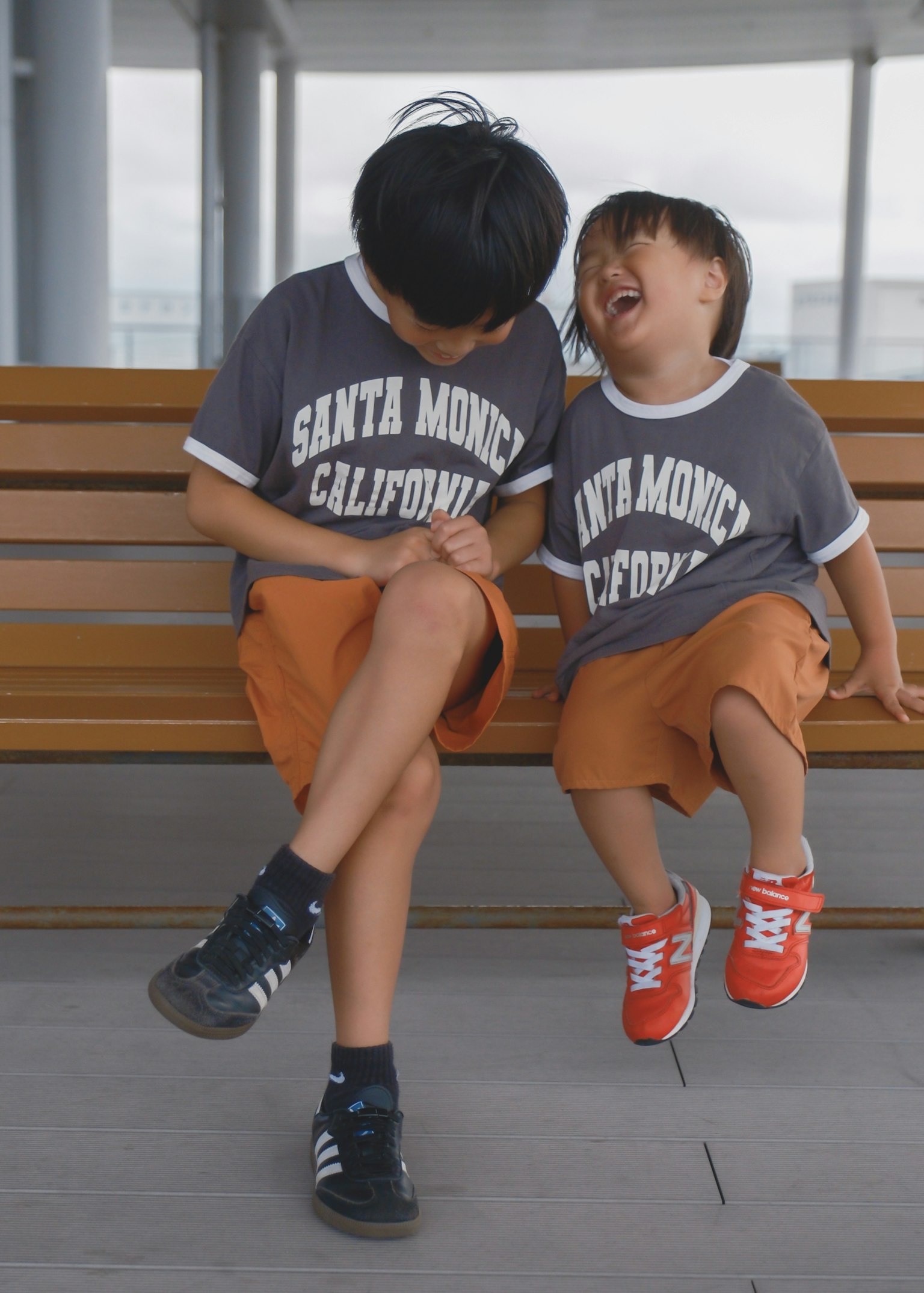 Dua anak duduk di bangku tersenyum mengenakan kaos Santa Monica California