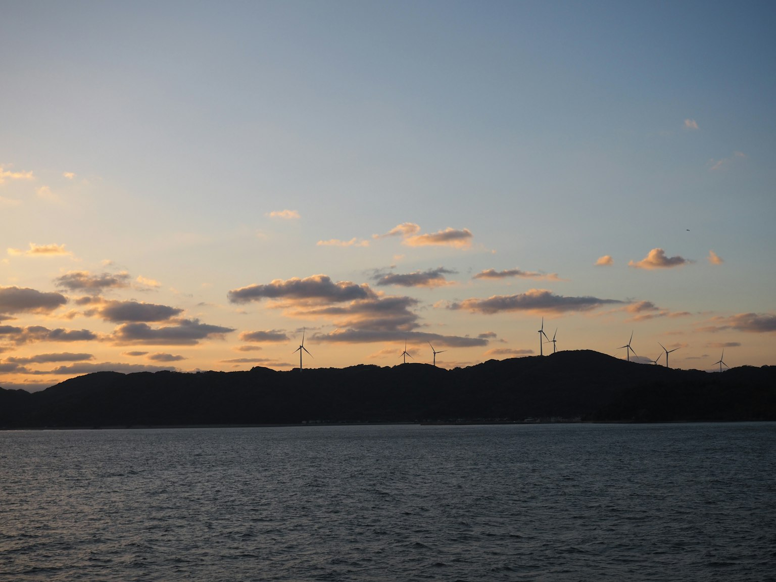 日落時分海面景色，遠處山丘上有風力發電機