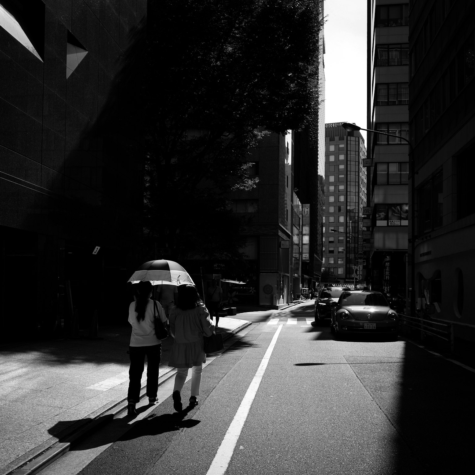 Dua wanita berjalan dengan payung di jalan kota dikelilingi gedung tinggi dan bayangan