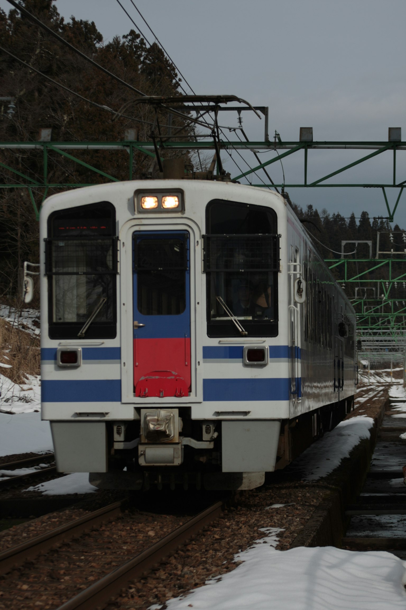 มุมมองด้านหน้าของรถไฟสีขาวที่มีแถบสีน้ำเงินบนรางที่มีหิมะ