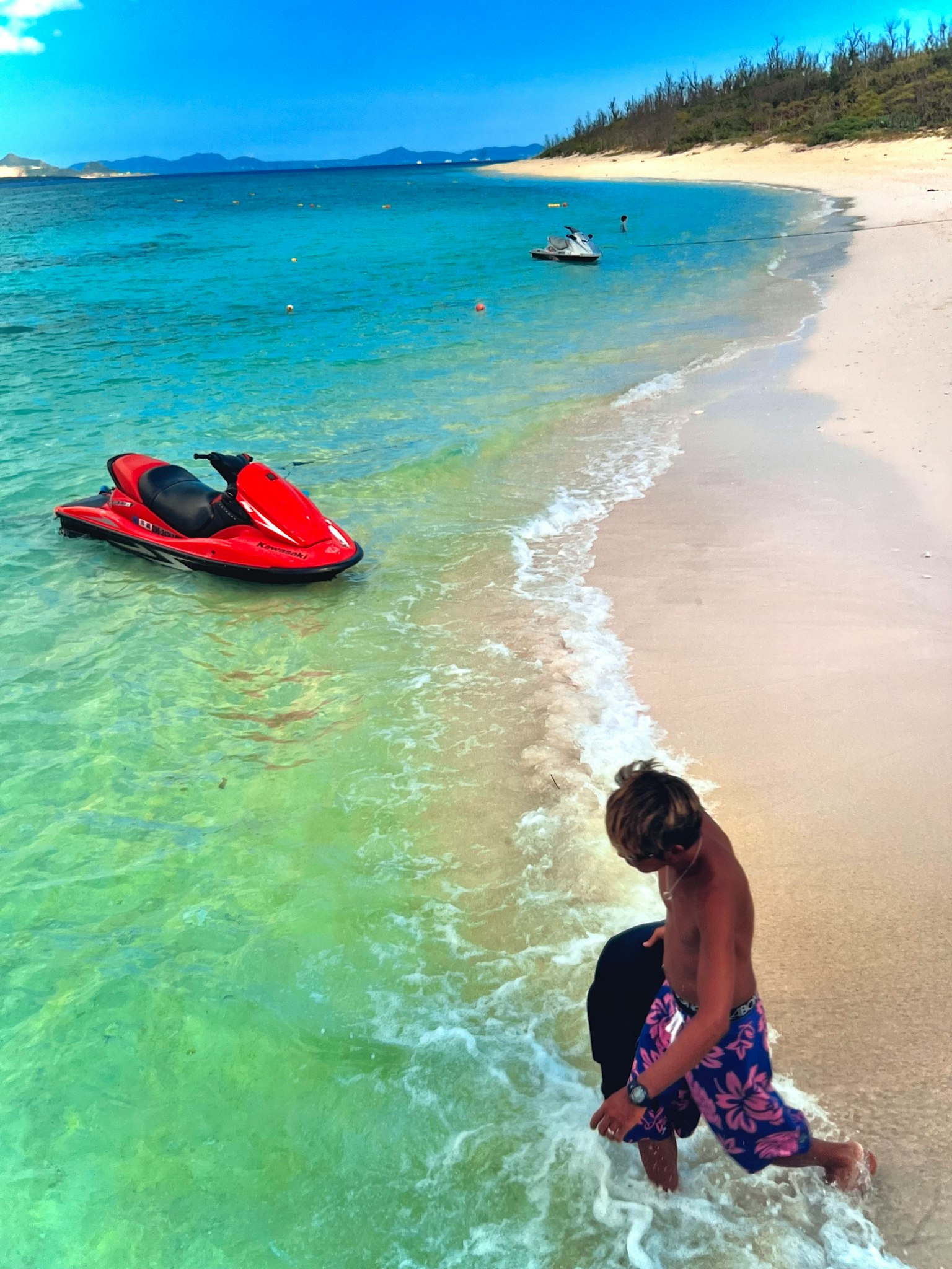 A woman walking along a beach with a jet ski in the clear water