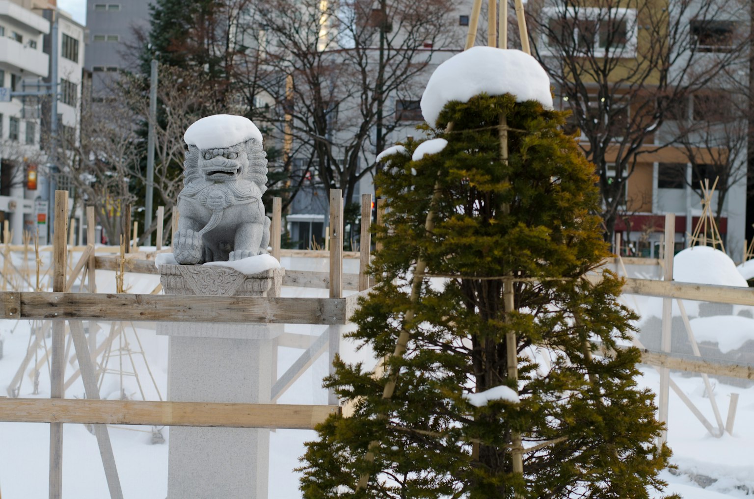 Statue en pierre d'un lion-chien recouverte de neige avec un arbre à proximité