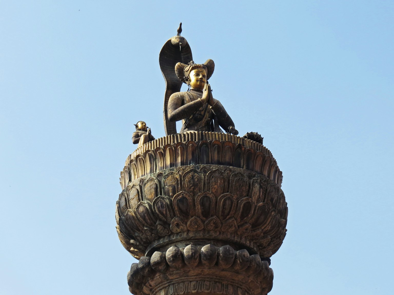 Statua di una divinità con le mani in preghiera in cima a una colonna decorativa sotto un cielo blu chiaro
