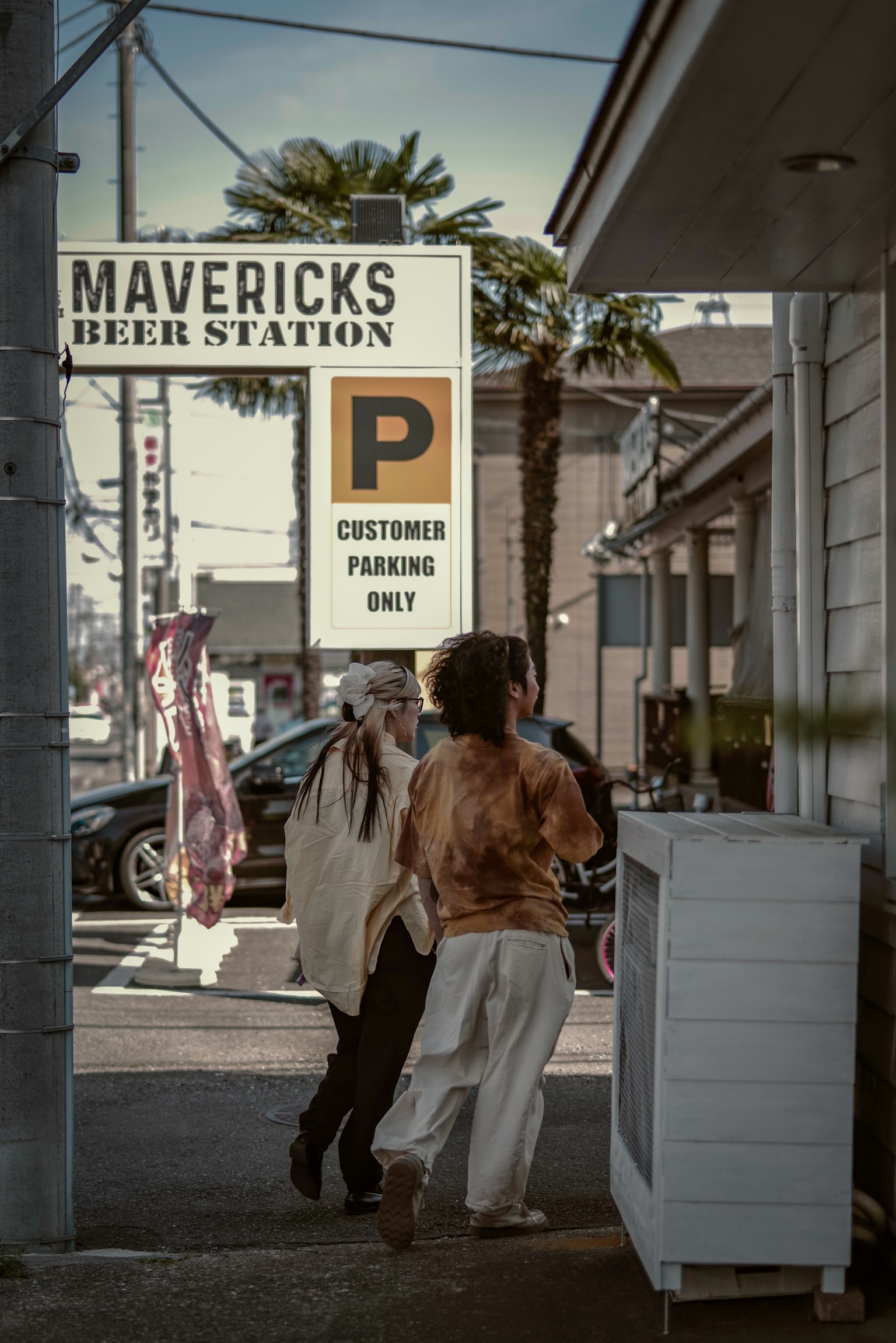 Zwei Personen gehen an dem Schild von Mavericks Beer Station auf einer sonnigen Straße vorbei