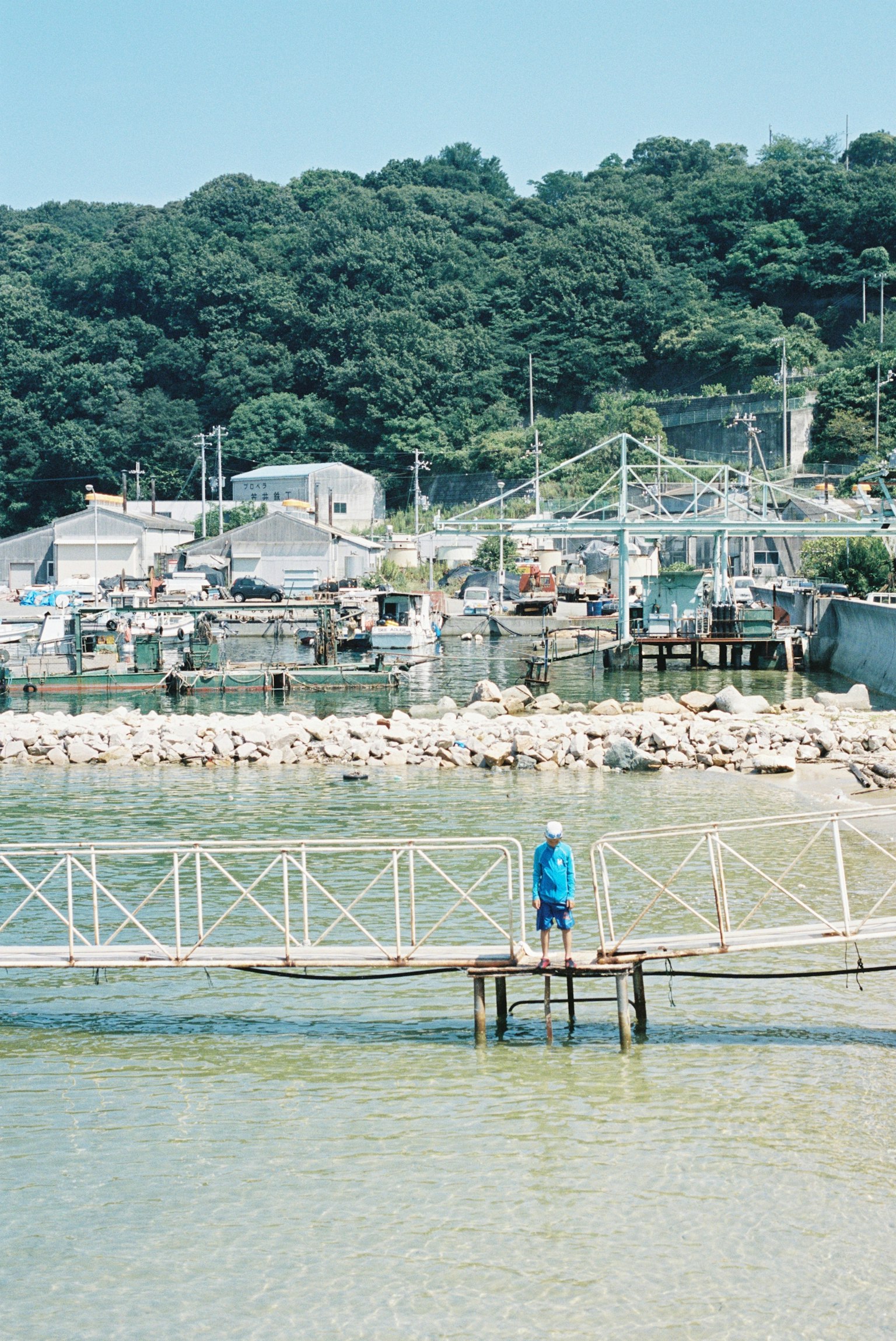 青い服を着た人が桟橋に立っている海の風景 背景には山と建物が見える