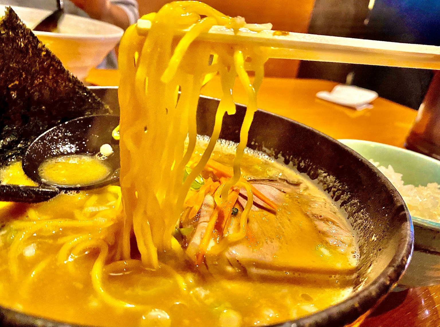 Ramen bowl with golden noodles and slices of meat held by chopsticks