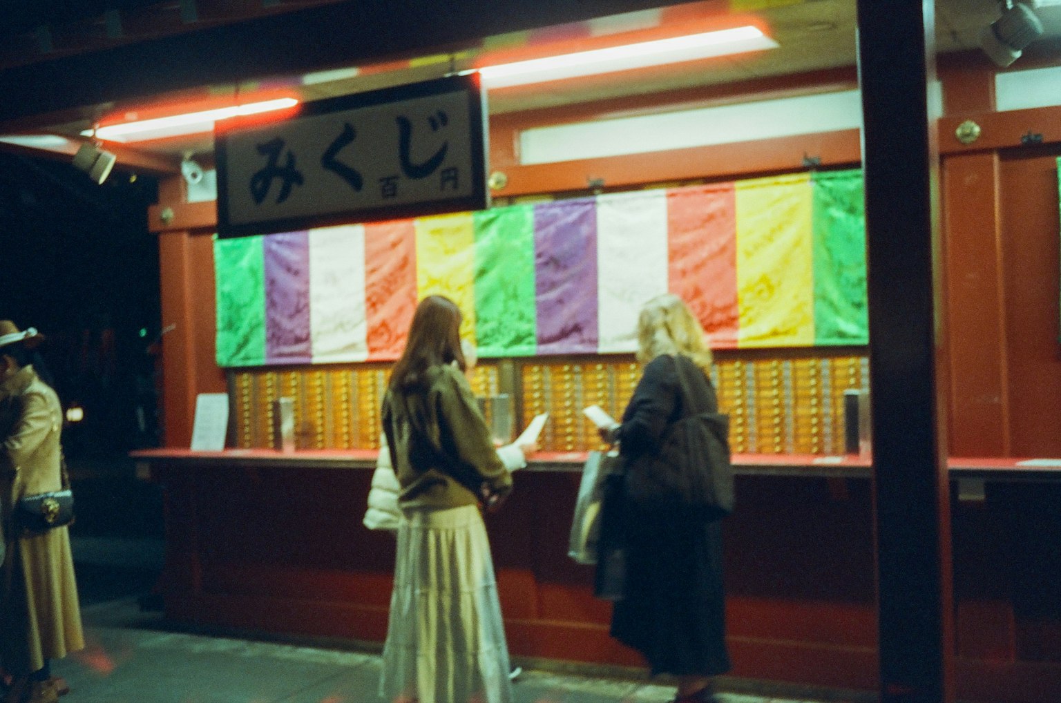 Mujeres sacando omikuji en un santuario por la noche Cortinas coloridas y cajas de madera visibles