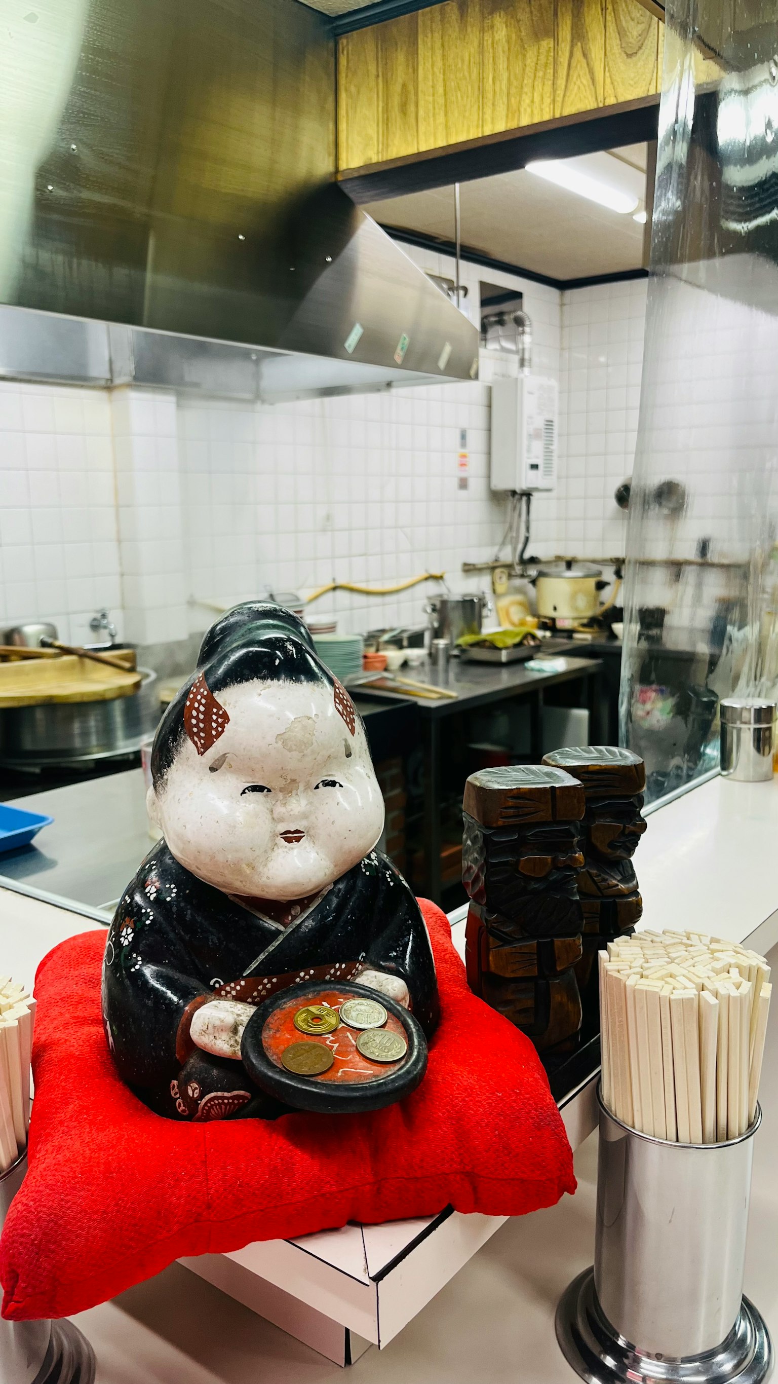 Una figura de gato con ropa tradicional sentada en un cojín rojo sosteniendo un tazón en el interior de un restaurante japonés