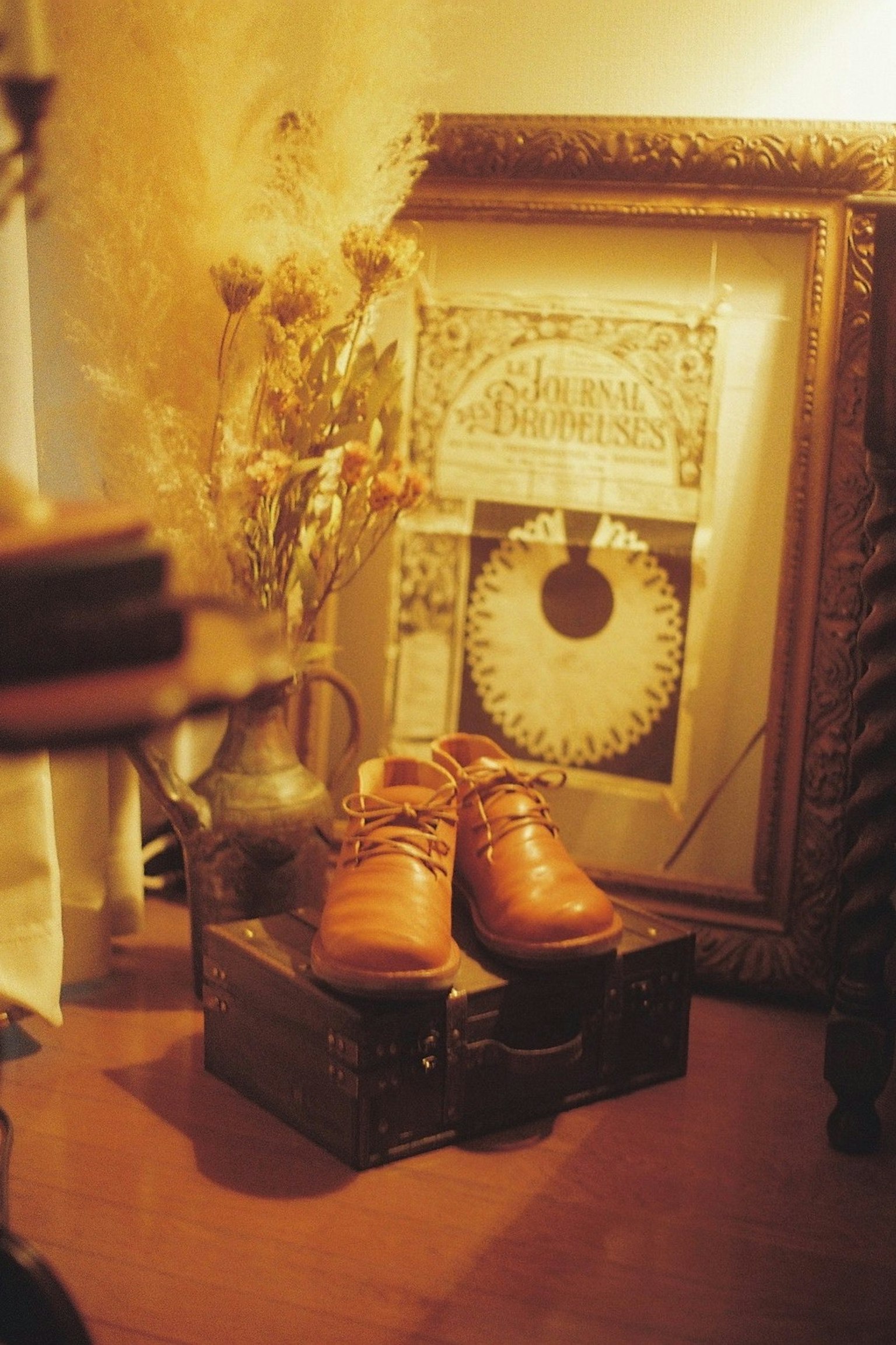 茶色の靴が木箱の上に置かれた静かな室内のシーン 壁に飾られたアートフレームとドライフラワーの花瓶が背景にある