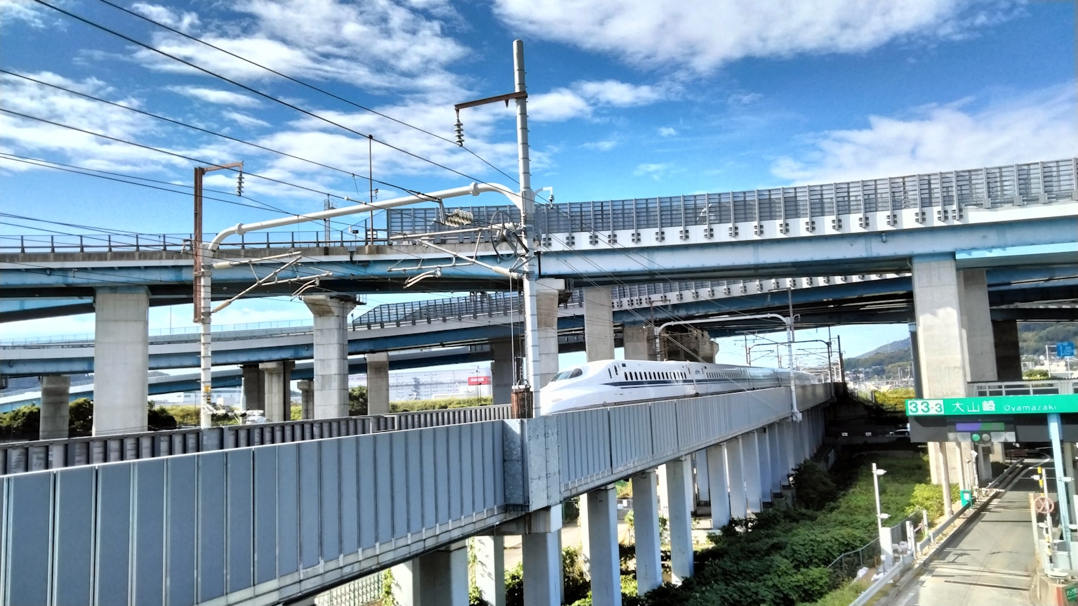 Shinkansen chạy dưới đường cao tốc với bầu trời xanh và mây