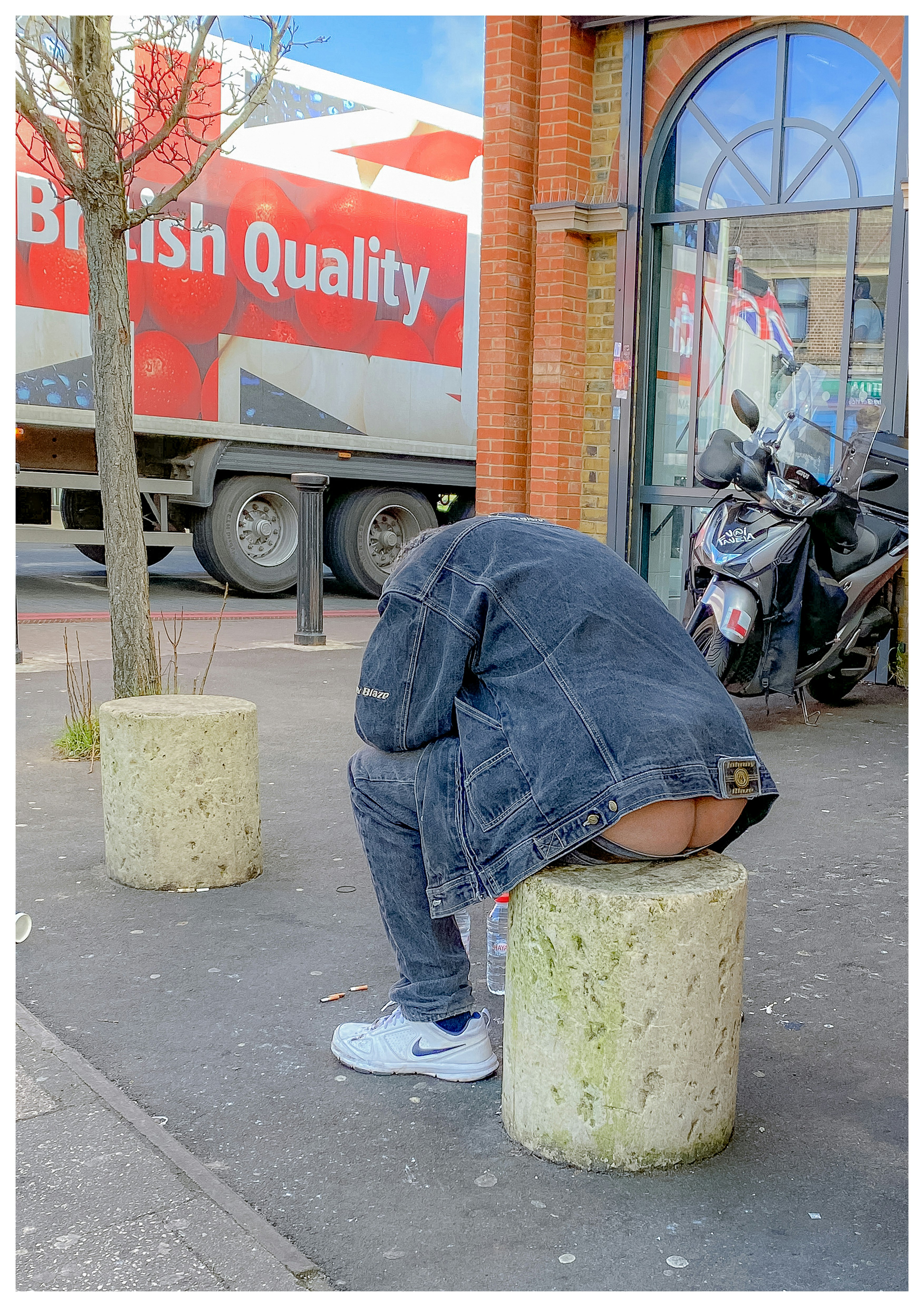 Eine Person hockt auf der Straße vor einem Schild „British Quality my arse“ mit Motorrädern in der Nähe