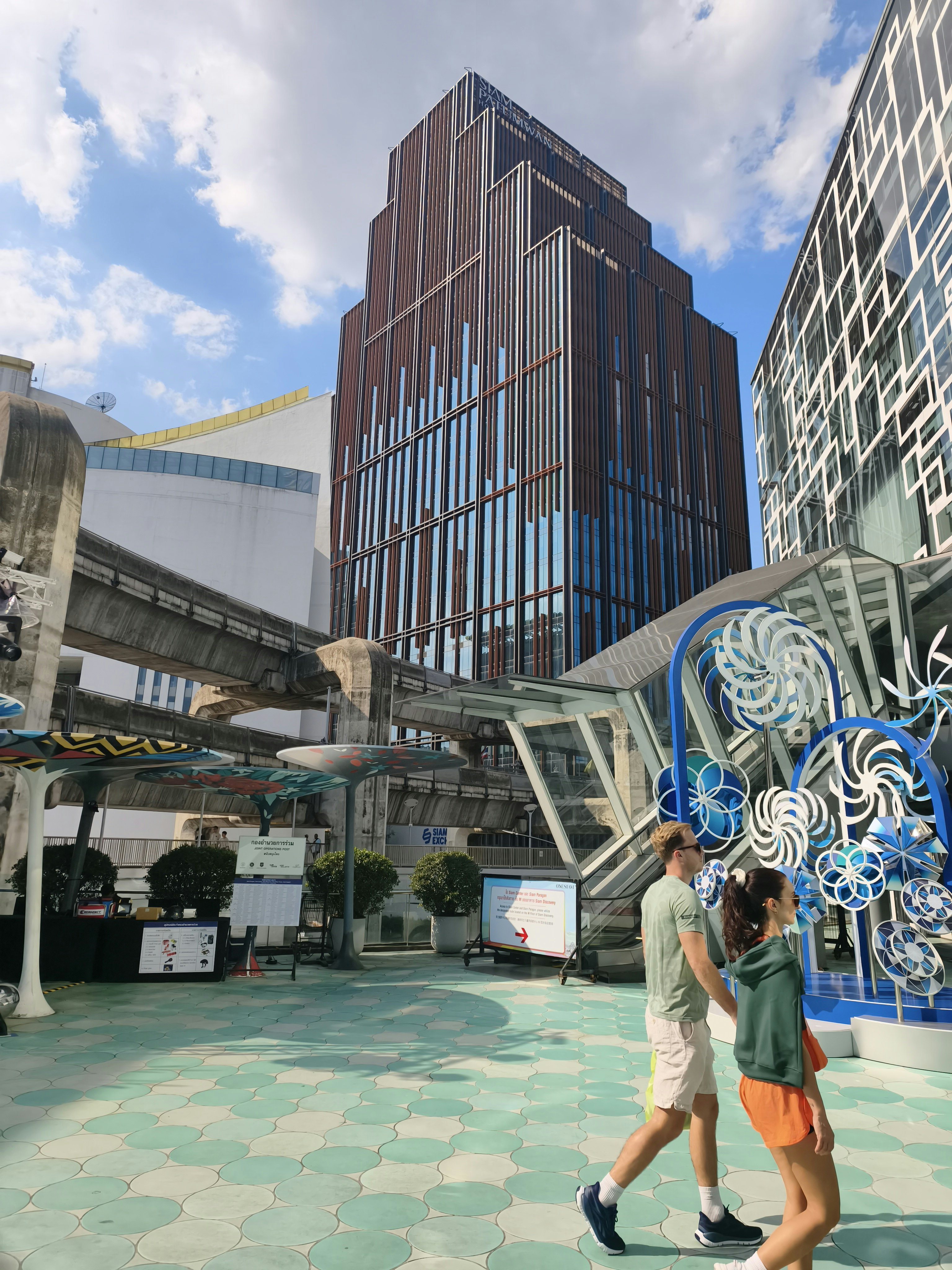 近未来的なビル群と屋外の歩道、金属のオブジェが並ぶ広場