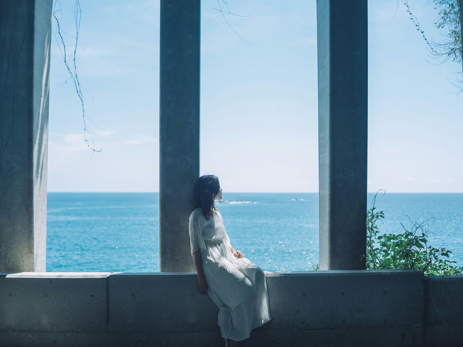 穿着白色裙子的女性坐在混凝土柱子之间，背景是蓝色的海洋