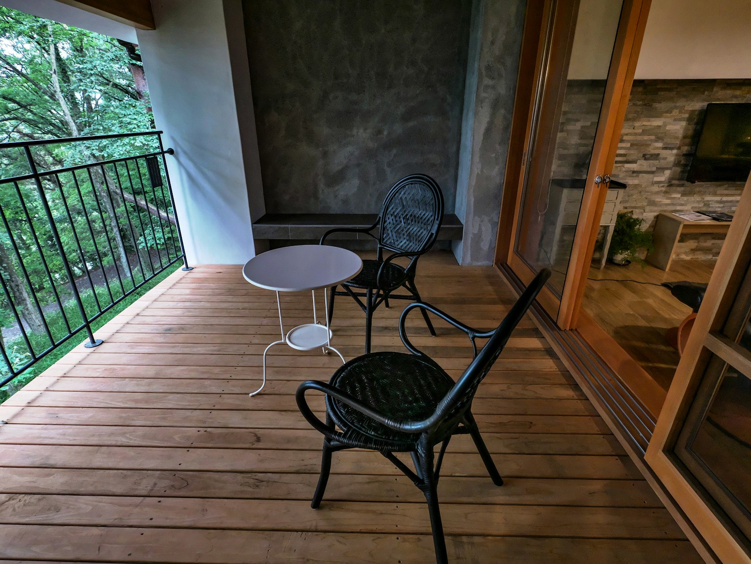 Ein Balkon mit Holzdeck, ausgestattet mit einem weißen Tisch und einem schwarzen Stuhl mit Blick auf das Grün