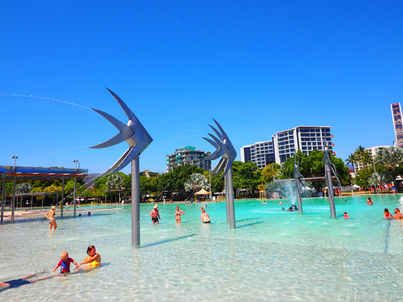 Bambini che giocano in un parco acquatico con sculture di pesci metallici e un cielo blu chiaro