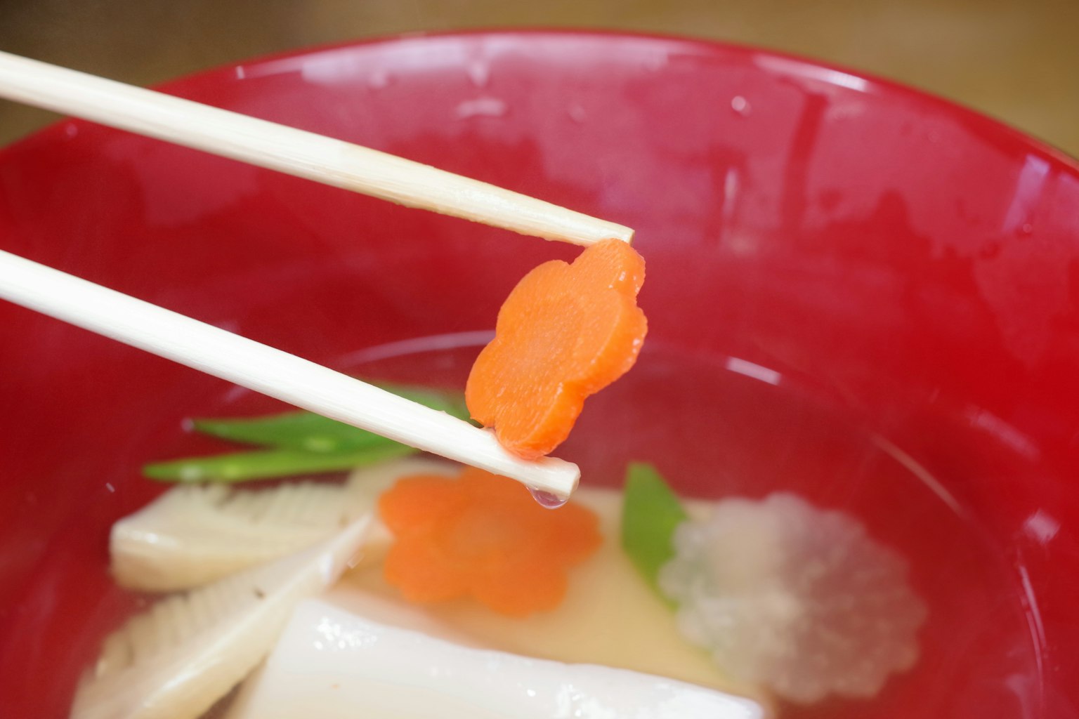 Carota a forma di fiore tenuta con le bacchette sopra una ciotola rossa di zuppa con verdure