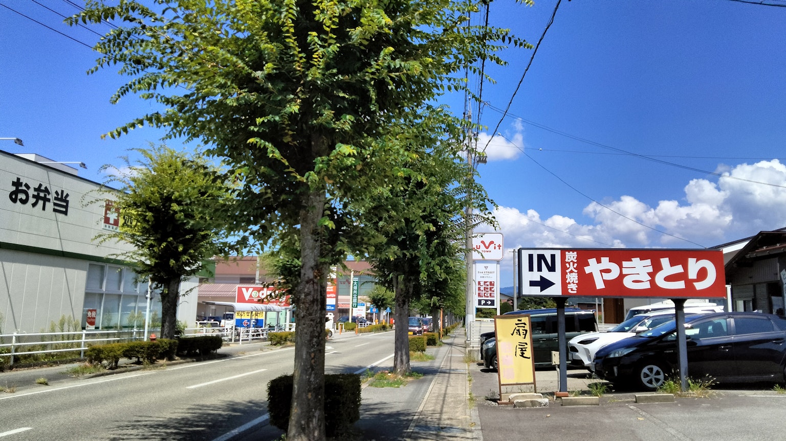 Pemandangan jalan dengan pepohonan dan bangunan komersial di bawah langit biru dengan tanda yakitori yang mencolok