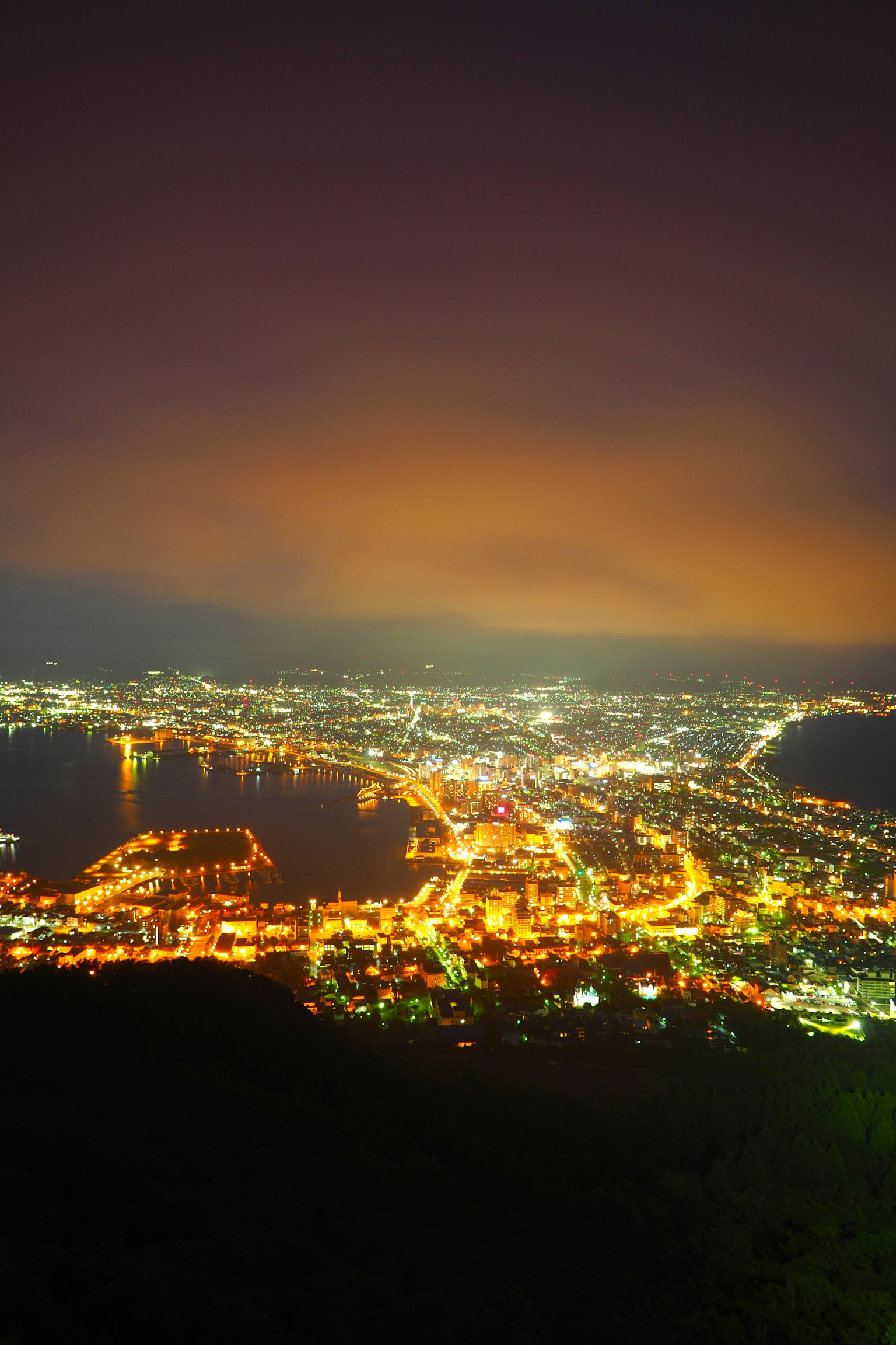Vista nocturna de una ciudad portuaria con luces brillantes y silueta de montaña