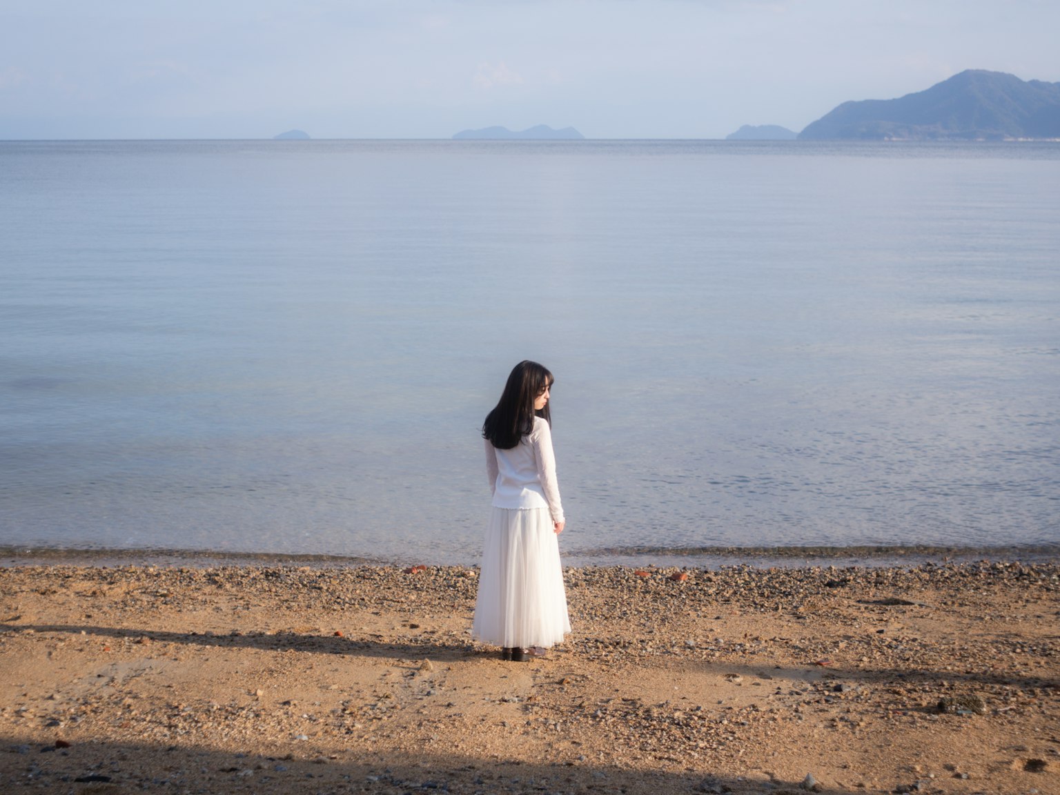 하얀 드레스를 입은 여성이 잔잔한 해변에 서서 바다를 바라보고 있다