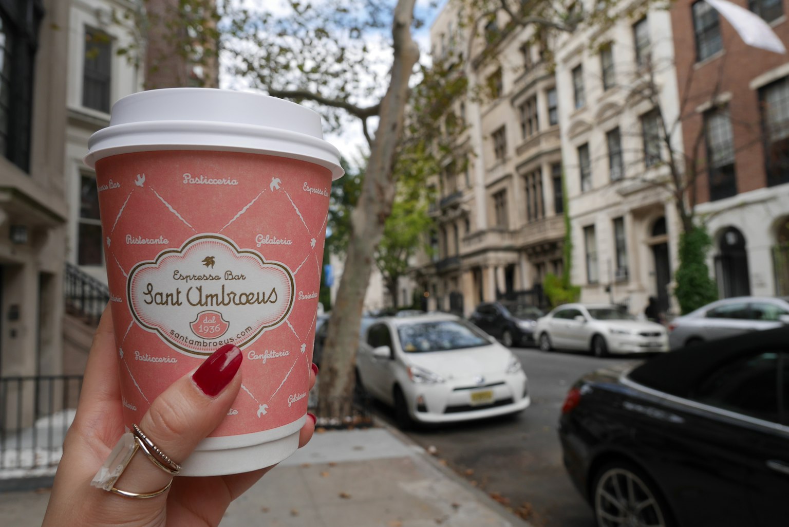 Una taza de café rosa sostenida en la mano con edificios al fondo