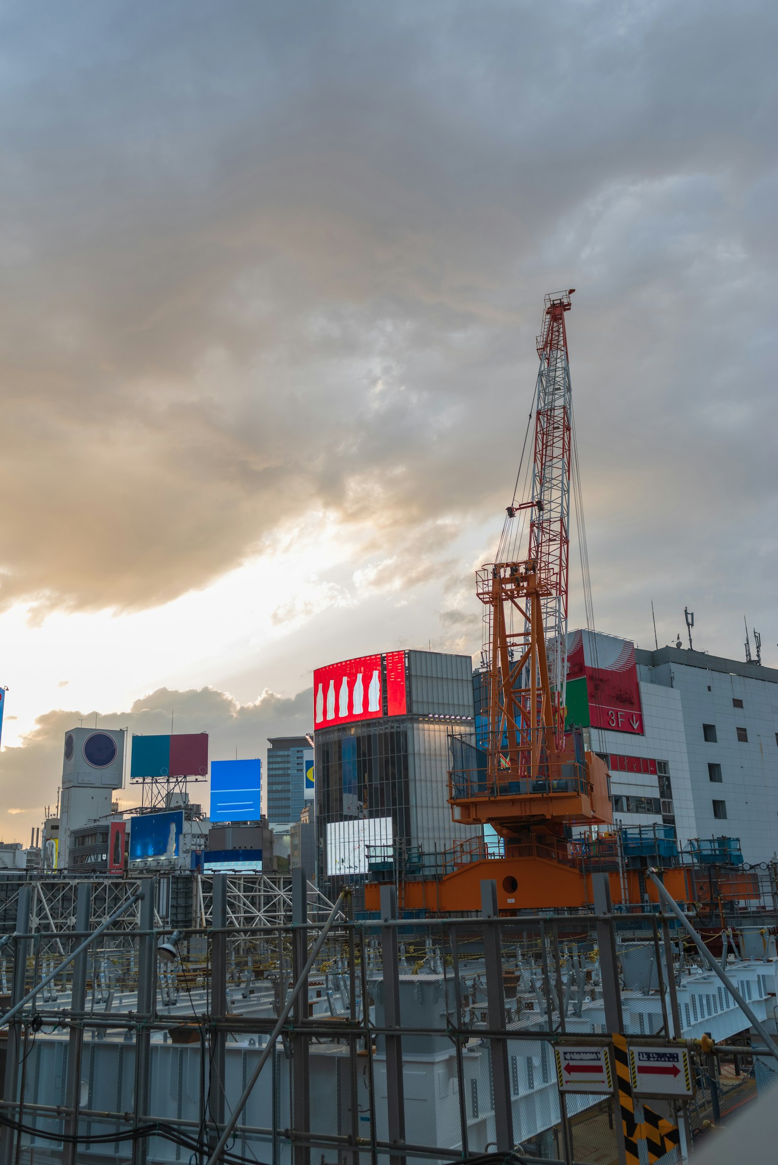 Sitio de construcción con grúa y letreros de edificios bajo un cielo al atardecer