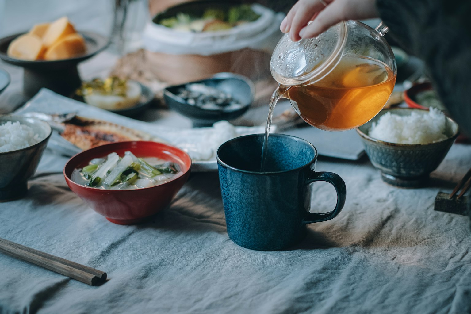 一只手将茶倒入蓝色杯子，桌上摆放着各种菜肴