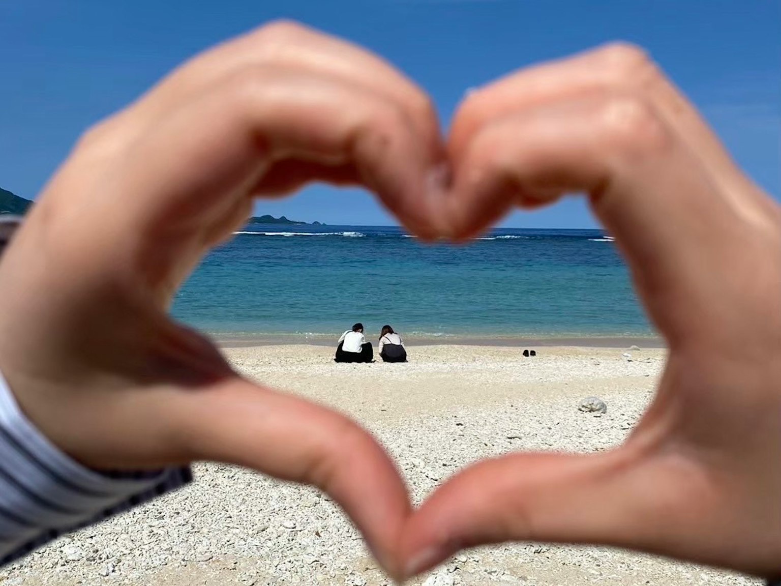 Tangan membentuk bentuk hati dengan pemandangan pantai dan dua orang duduk di latar belakang