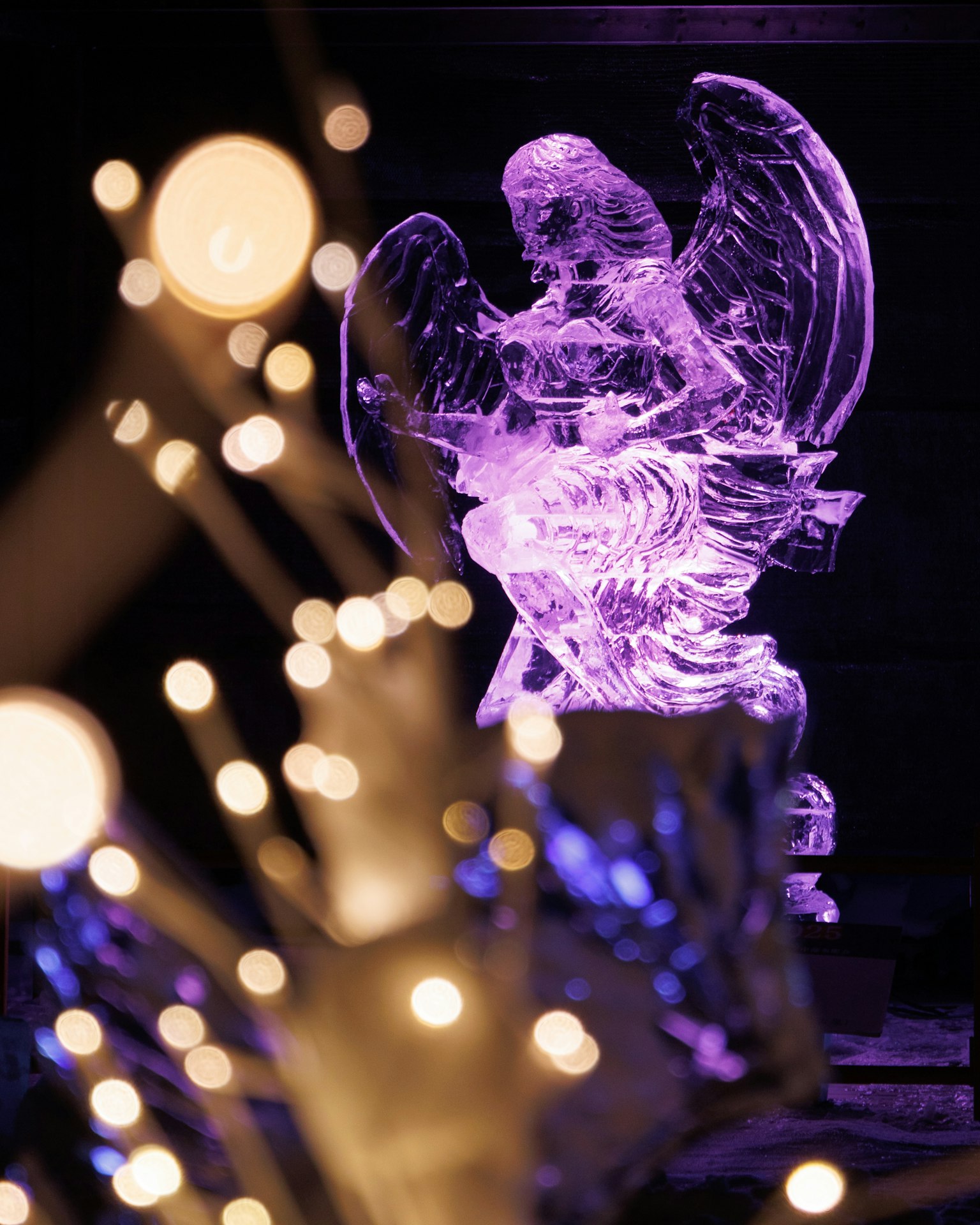 Sculpture de glace en forme d'ange illuminée par une lumière violette avec des lumières floues au premier plan