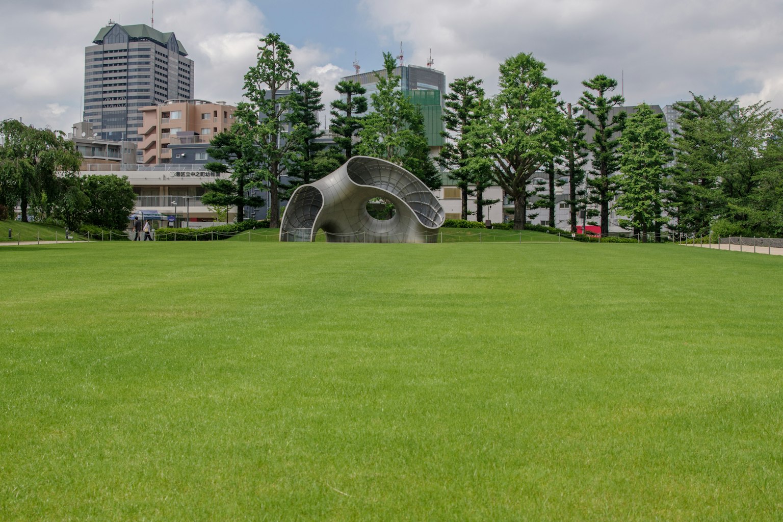 Patung modern di atas rumput hijau dengan gedung pencakar langit di latar belakang