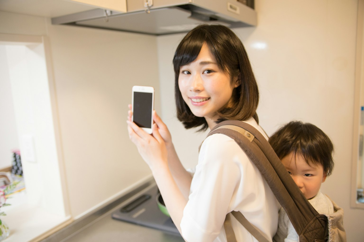 Femme tenant un smartphone dans une cuisine avec un enfant sur son dos
