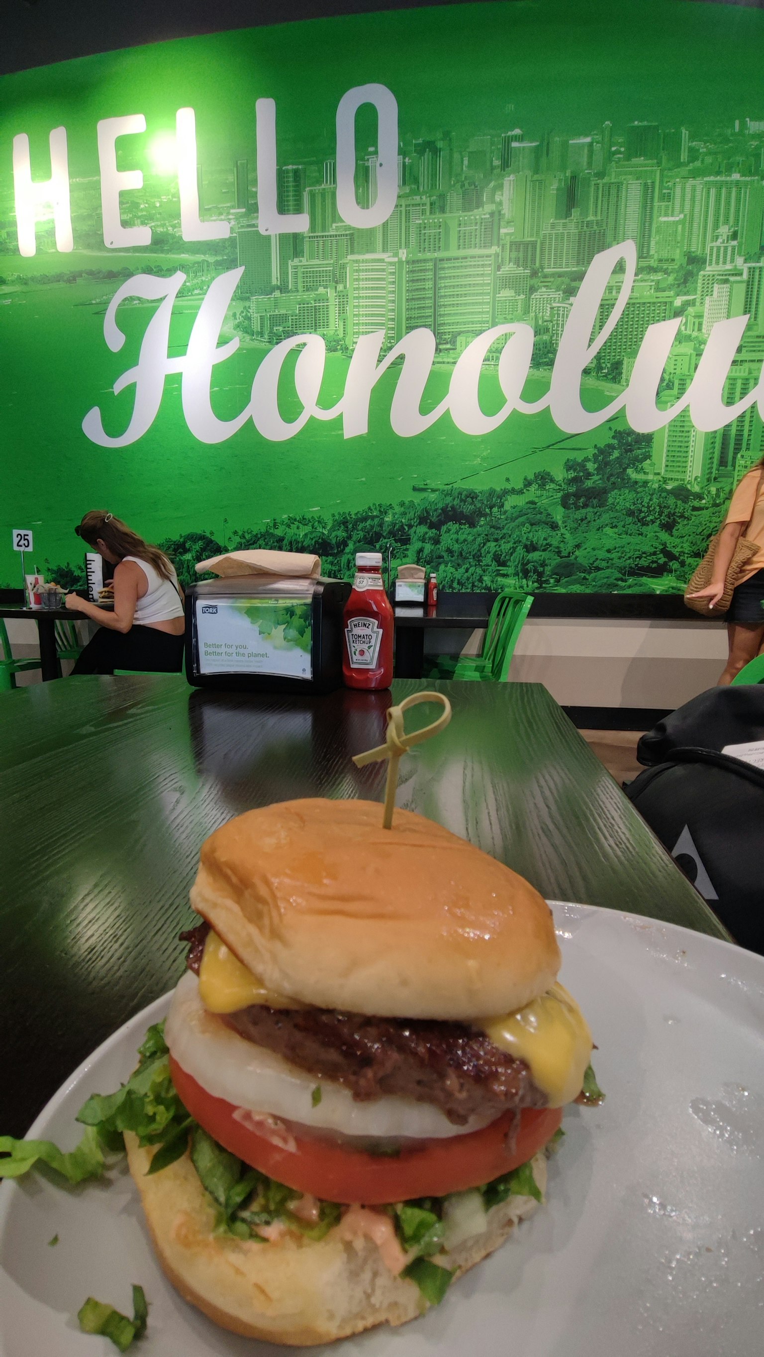 Una hamburguesa en un plato frente a una pared verde con las palabras Hola Honolulu