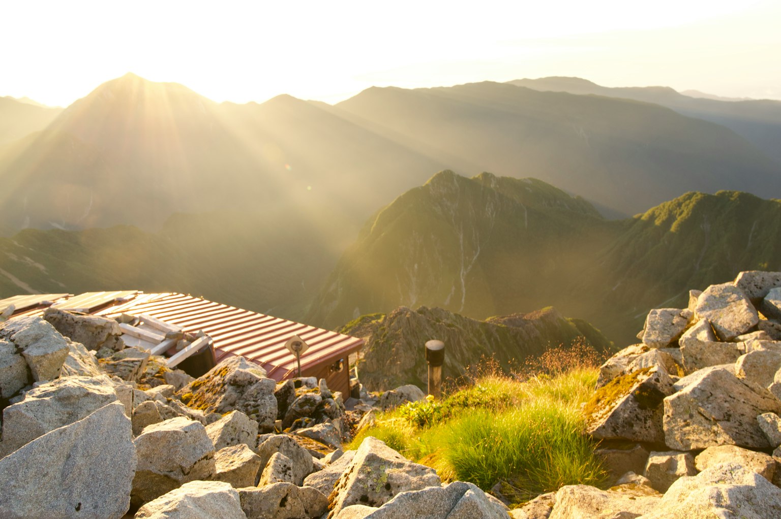 Vue panoramique depuis un sommet de montagne avec un cabanon entouré de rochers et d'herbe illuminé par le coucher de soleil