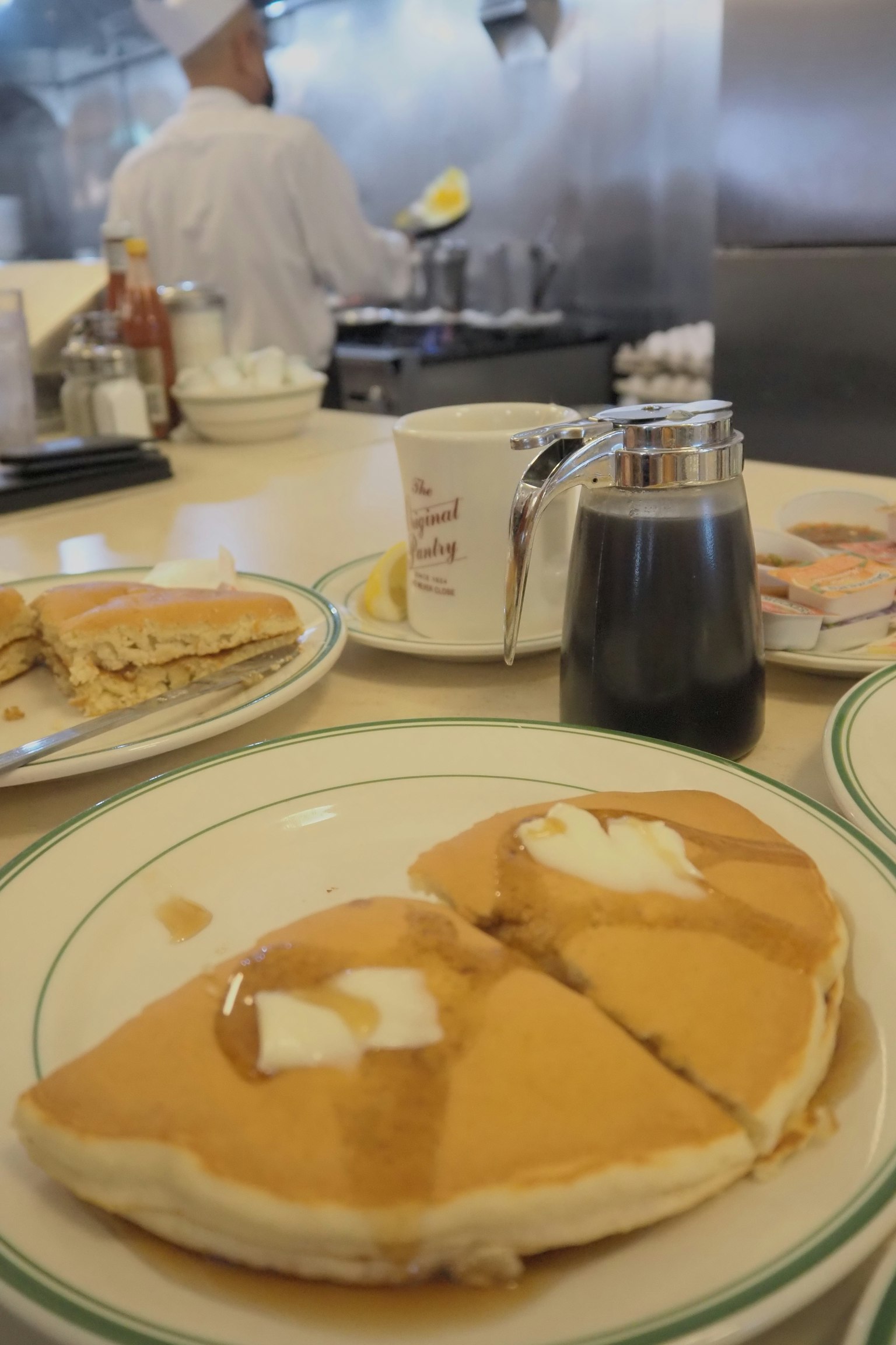 Cảnh bữa sáng với bánh pancake trên đĩa và một đầu bếp đang nấu ăn ở phía sau