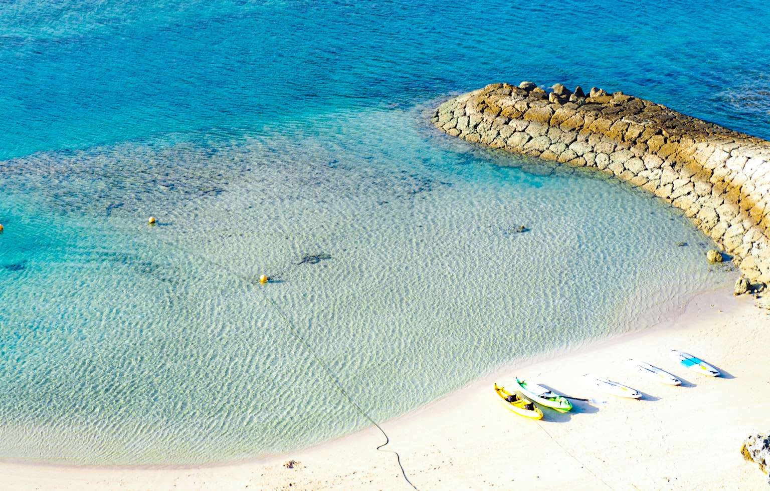 Vista aerea di una spiaggia con acqua blu chiara e sabbia, con un molo roccioso e barche colorate