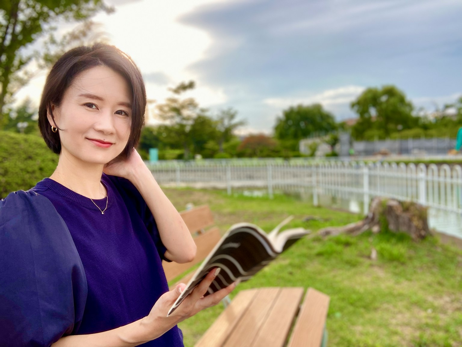 Người phụ nữ mỉm cười trong công viên cầm sách với nền xanh và bầu trời xanh