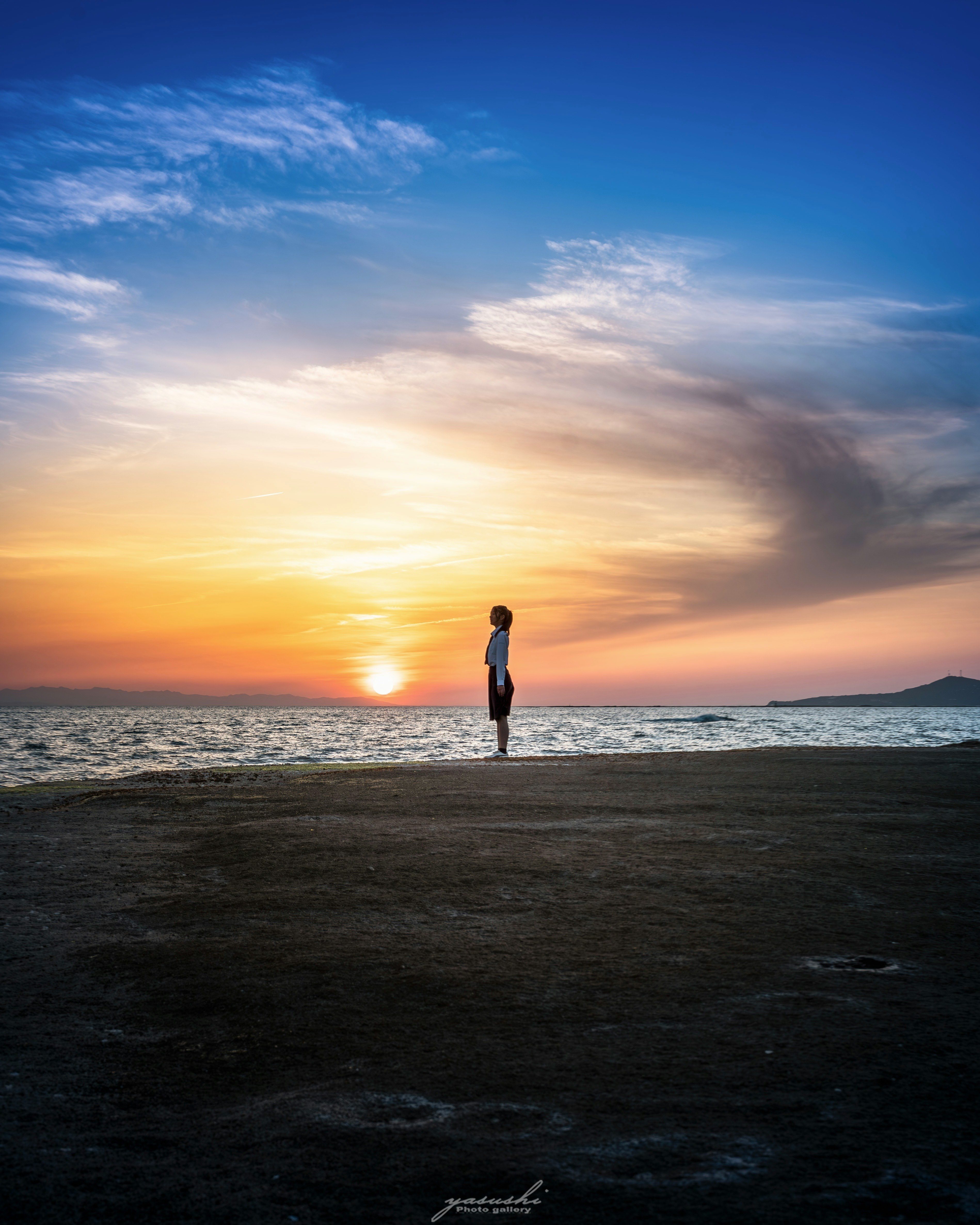 地平線に沈む夕陽を見つめる人物と海辺の風景