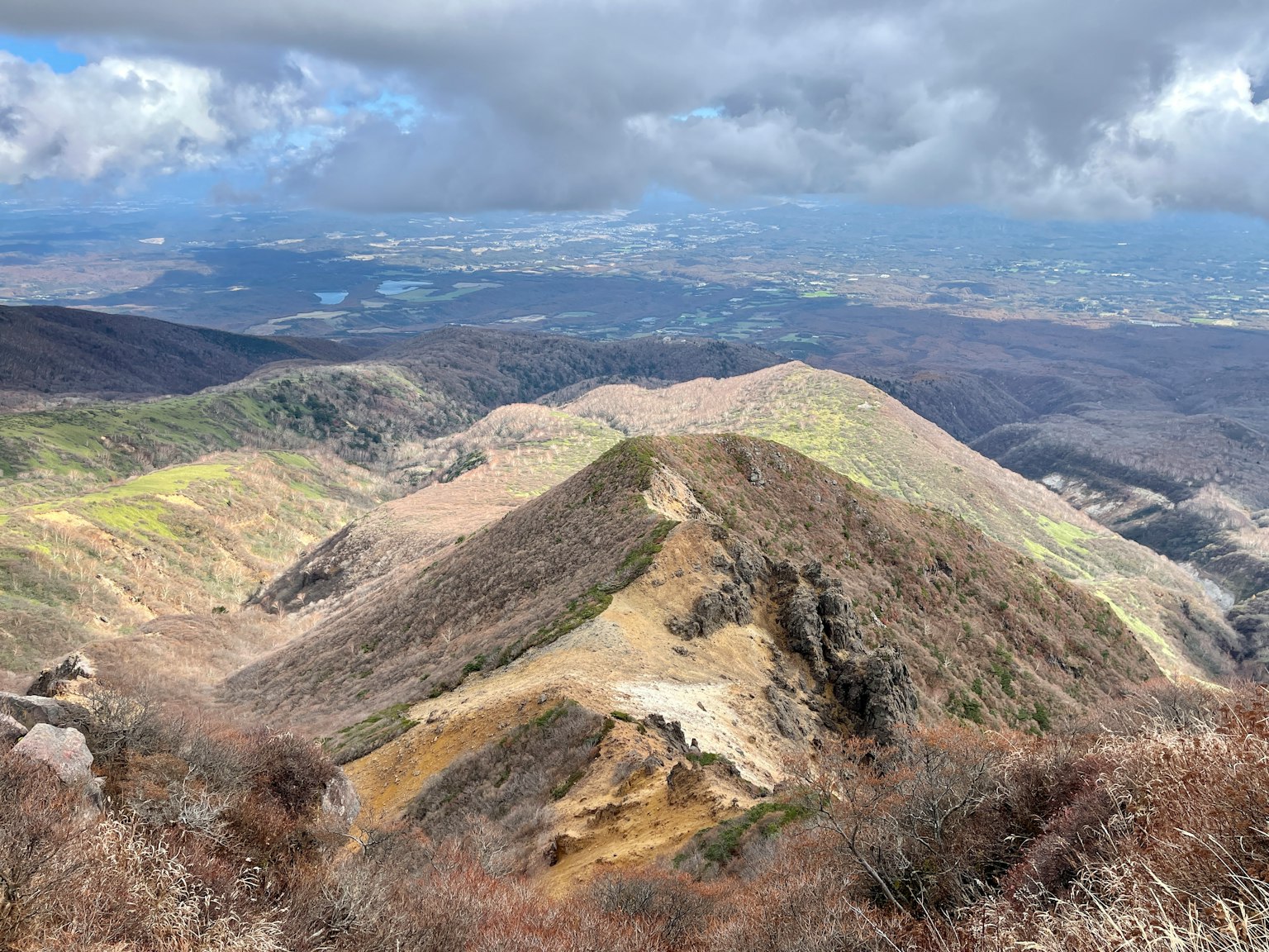 Vista escénica desde la cima de una montaña con pendientes verdes y marrones bajo un cielo nublado